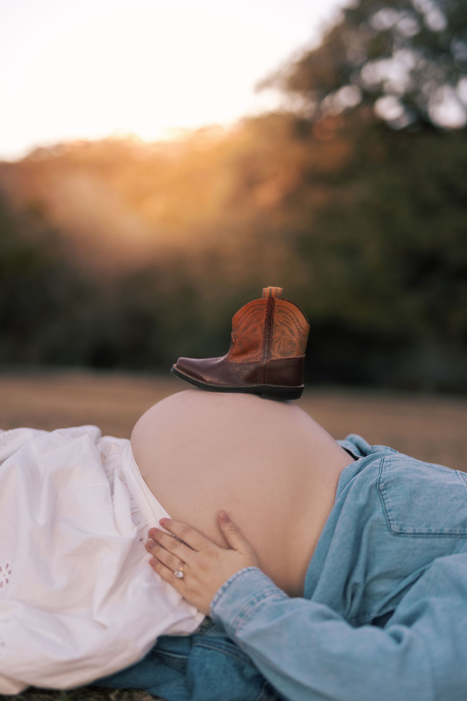 A pregnant woman in a denim shirt lies on her side with her hand resting on her baby bump during golden hour.