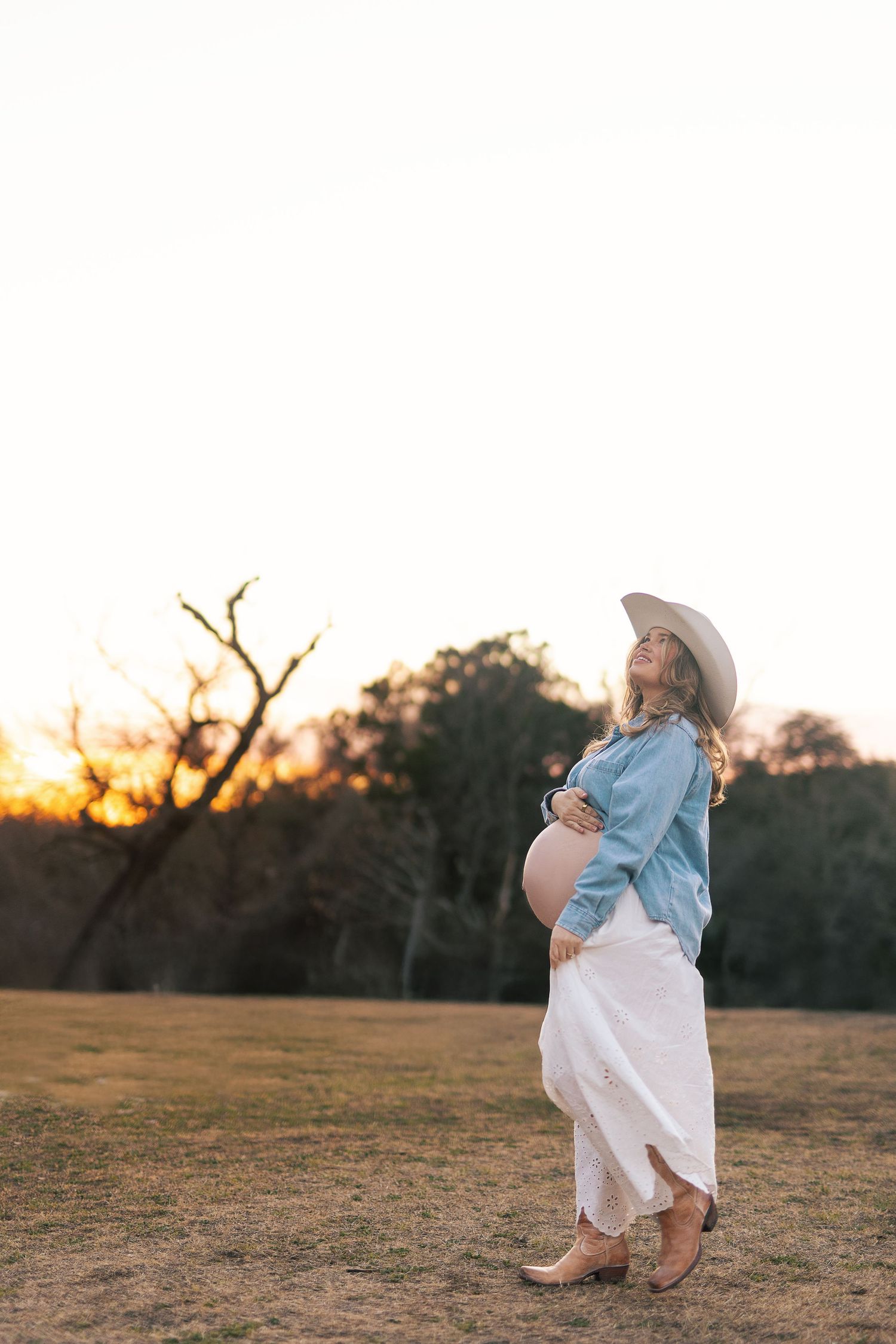 A person in a flowing white dress and blue top twirls gracefully in a grassy field at sunset.