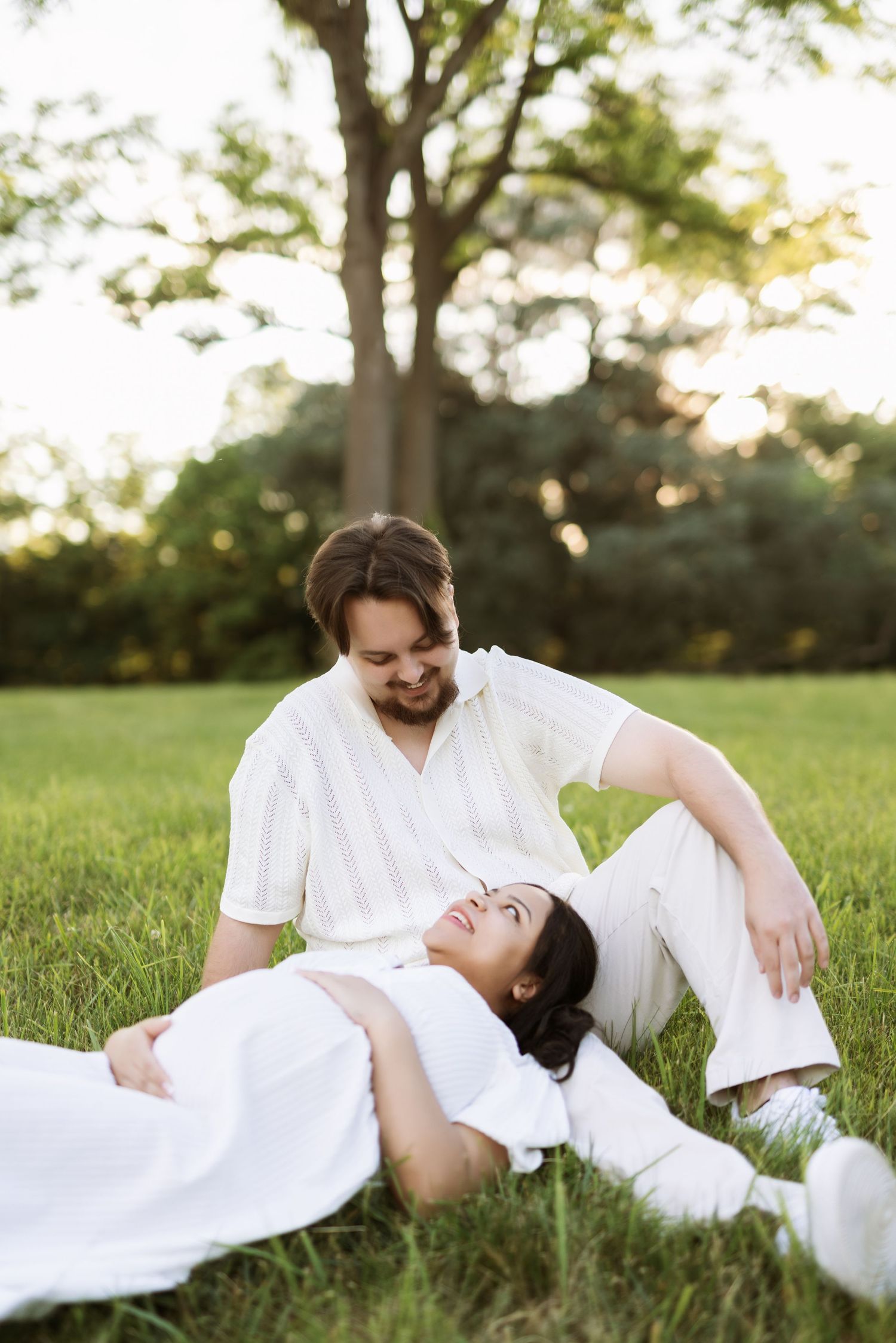 Couple in white clothing relaxing on grass during sunset photoshoot.