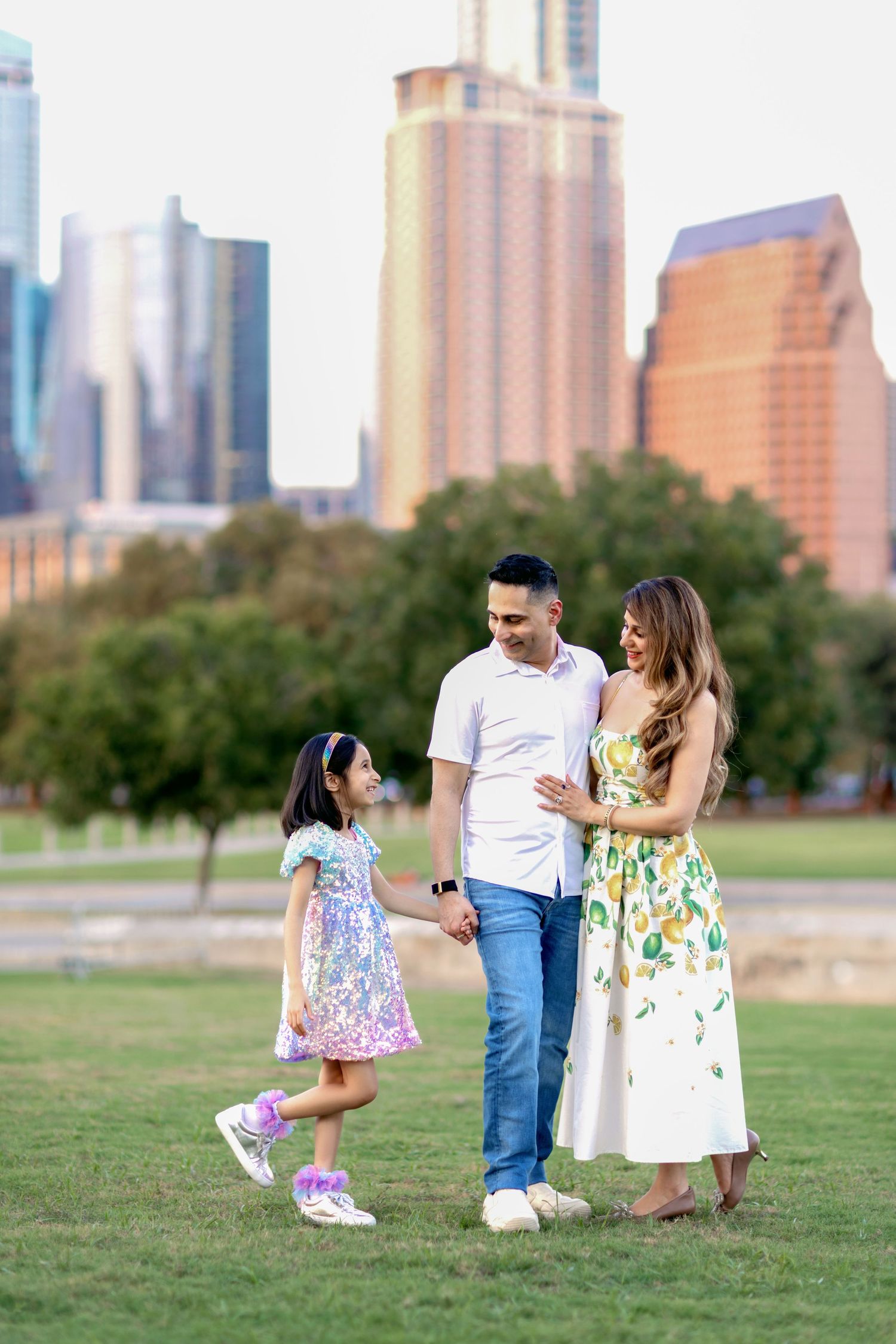 Family enjoying a playful moment together on a grassy field with city skyline in background.