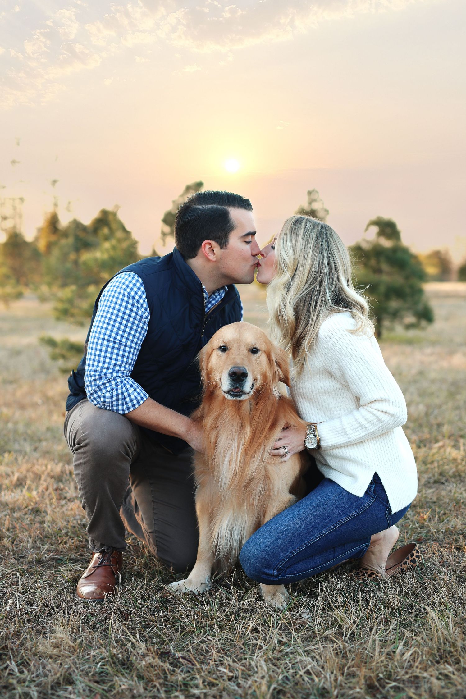 A happy Golden Retriever sits between a couple sharing a romantic moment at sunset.