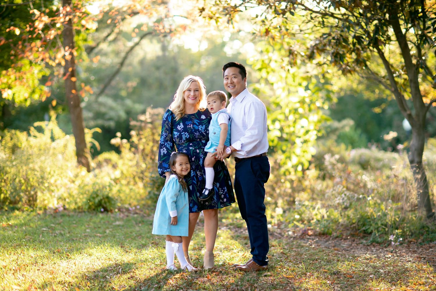 Family of three poses together in sunlit wooded area during golden hour.