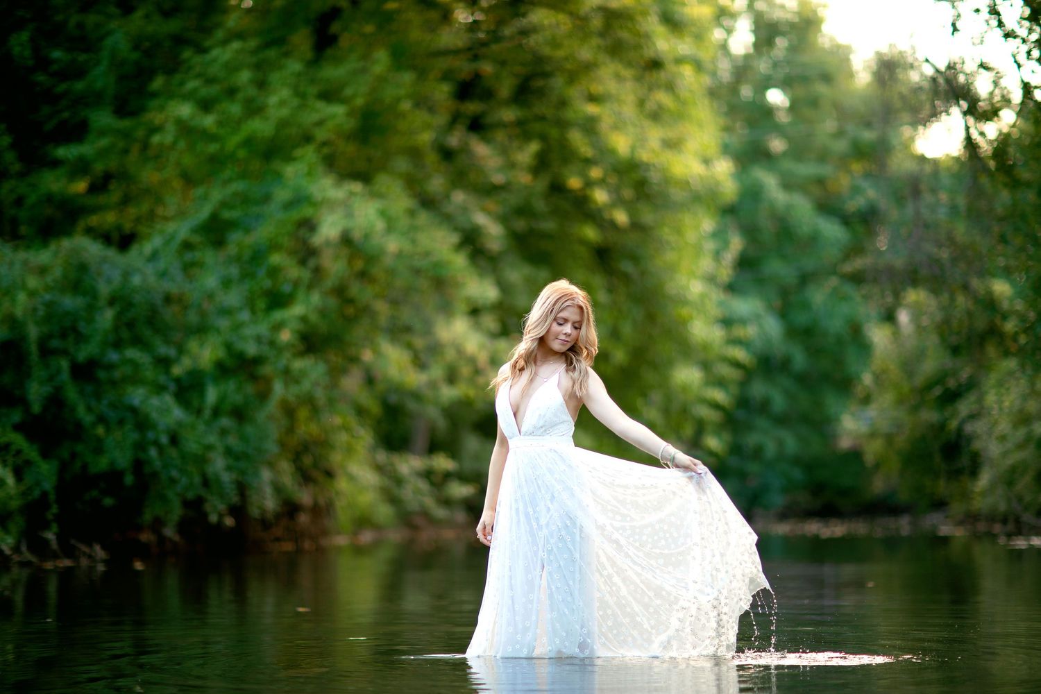 Woman in flowing white dress twirls gracefully by tranquil pond at sunset.