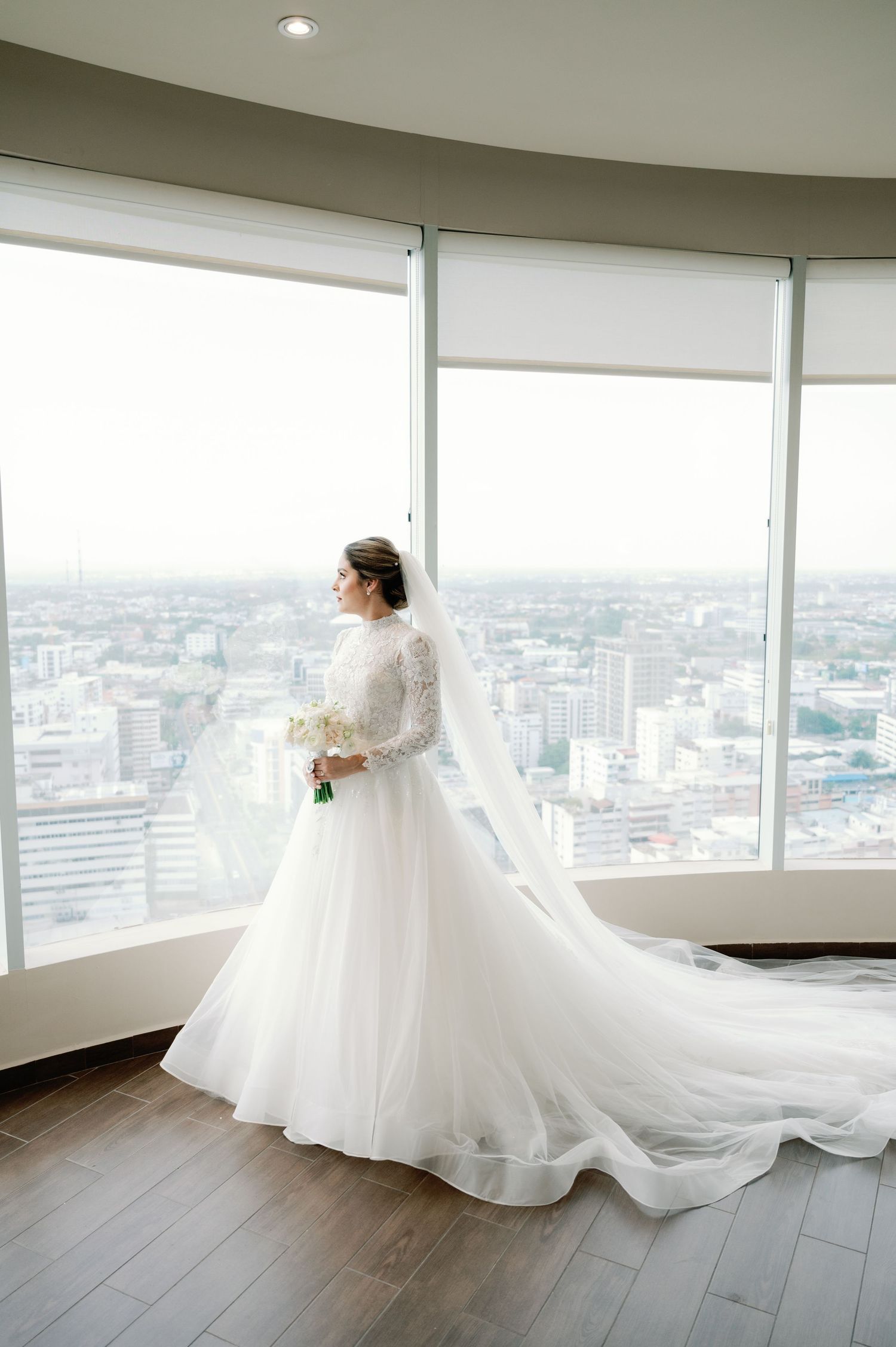 A flowing white wedding gown with a long train is showcased against floor-to-ceiling windows overlooking a cityscape.