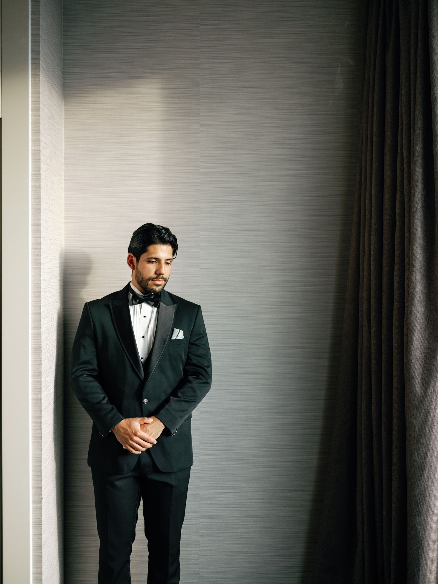 A groom in a classic black tuxedo poses thoughtfully against a modern grey wall with dramatic lighting.