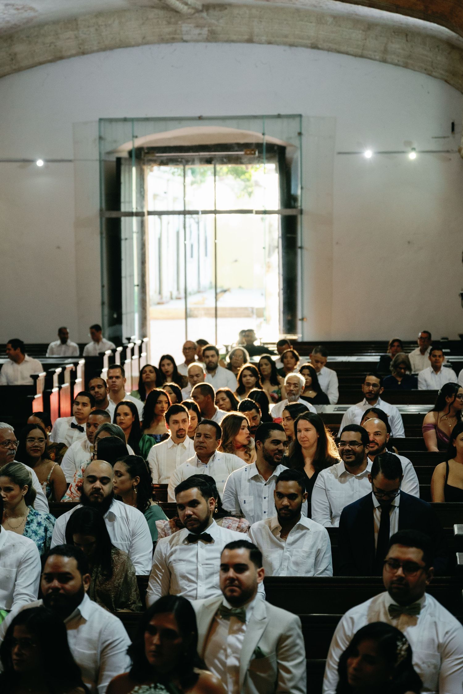 A large group of people wearing white shirts sits in rows of chairs in what appears to be an auditorium setting.