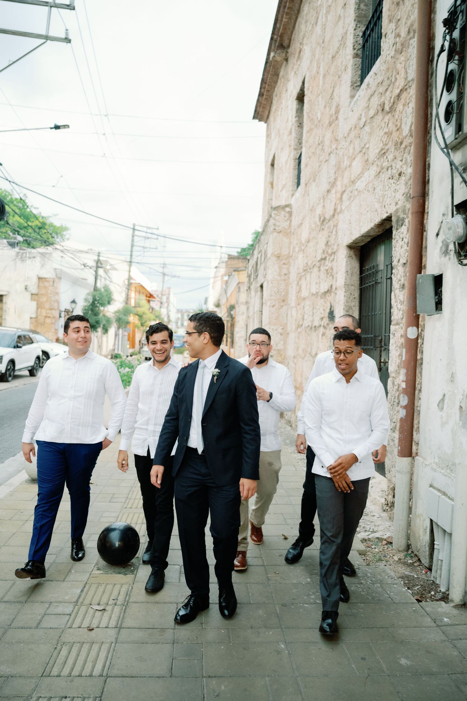 Group walking along a stone wall exterior in formal attire on a sunny day.