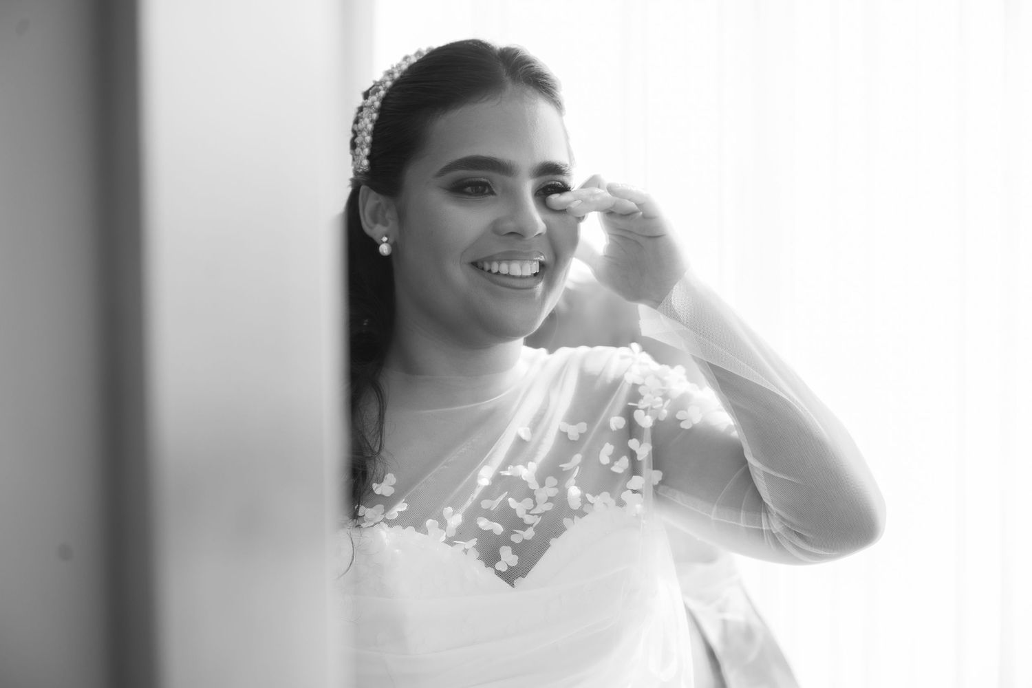 A bride in an elegant white lace gown stands in front of a bright window with natural light streaming in.