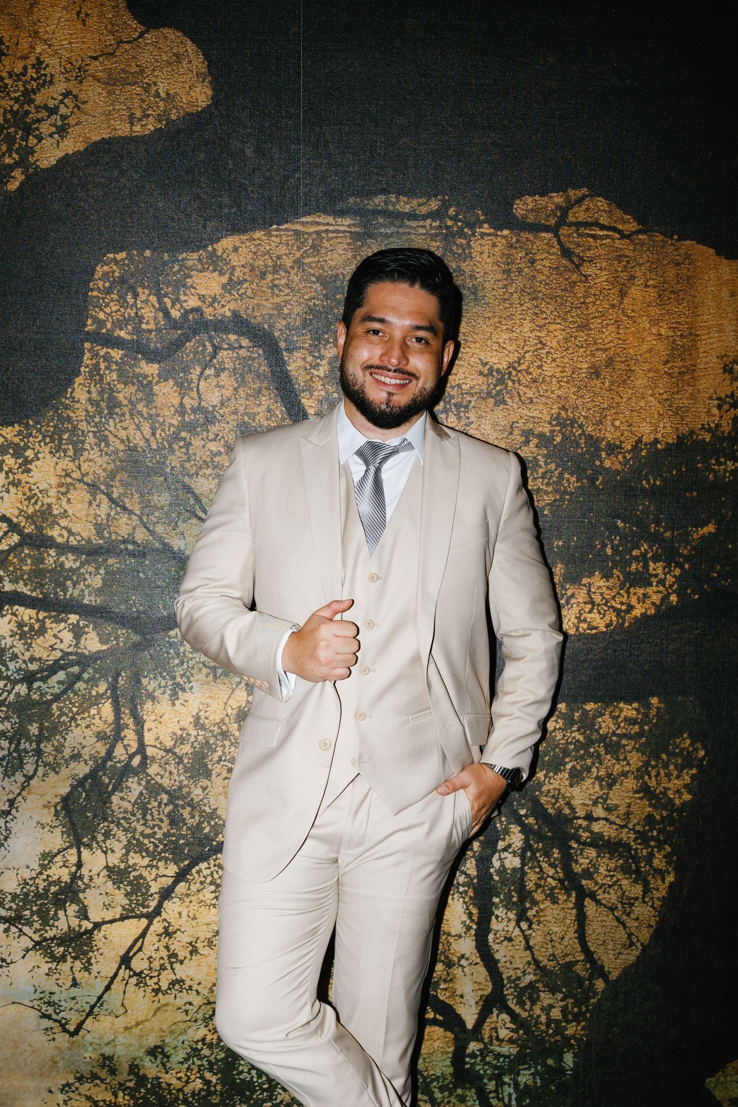 A person in a light beige suit poses against a dark textured wall with gold accents.