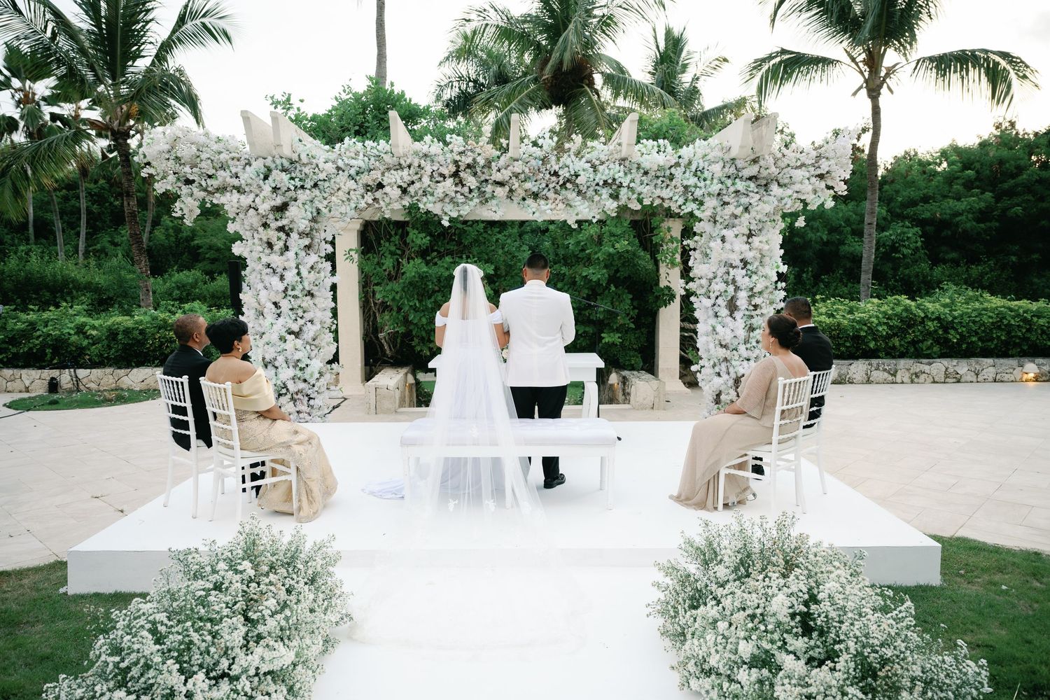 Elegant outdoor wedding ceremony setup with white floral arch and palm trees in tropical garden setting.
