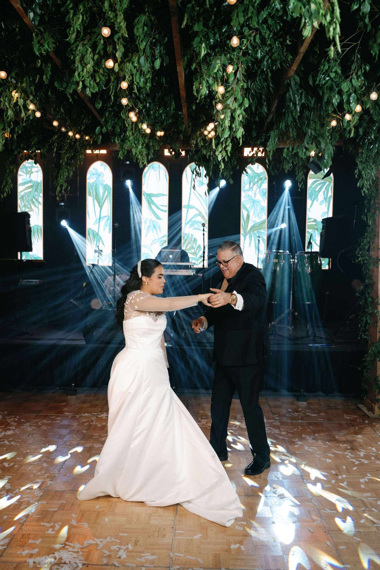 A couple dances together under hanging greenery and string lights on a brick patio.