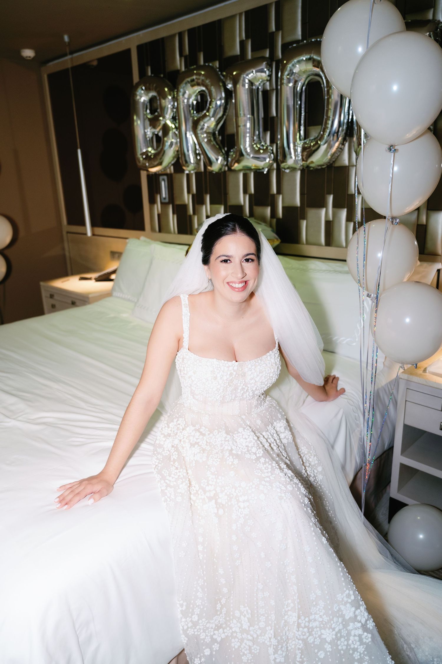 A bride in a sparkly ballgown poses with silver letter balloons and white decorations.