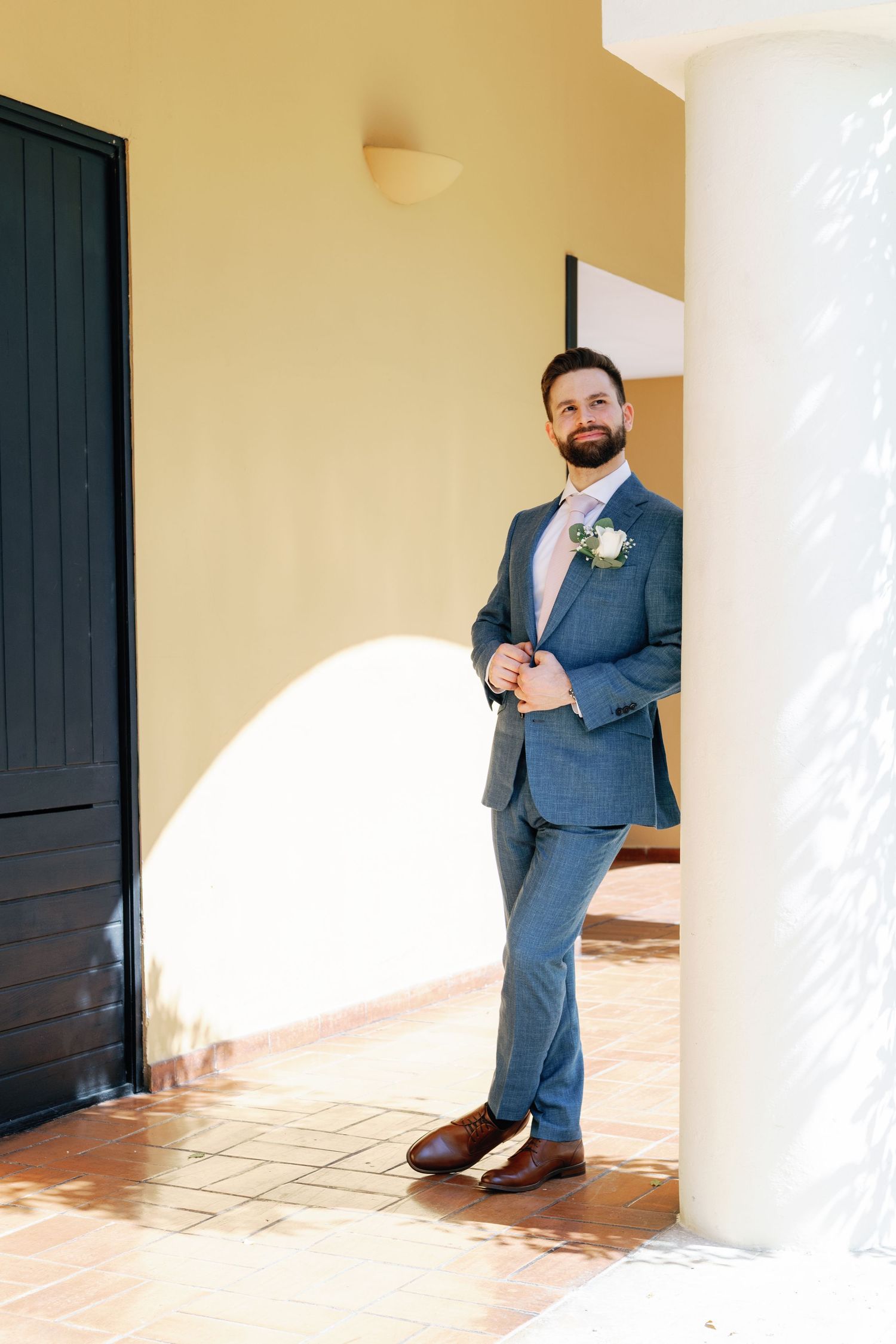 A person in a blue suit and boutonniere stands in a sunlit archway with terracotta tile flooring.