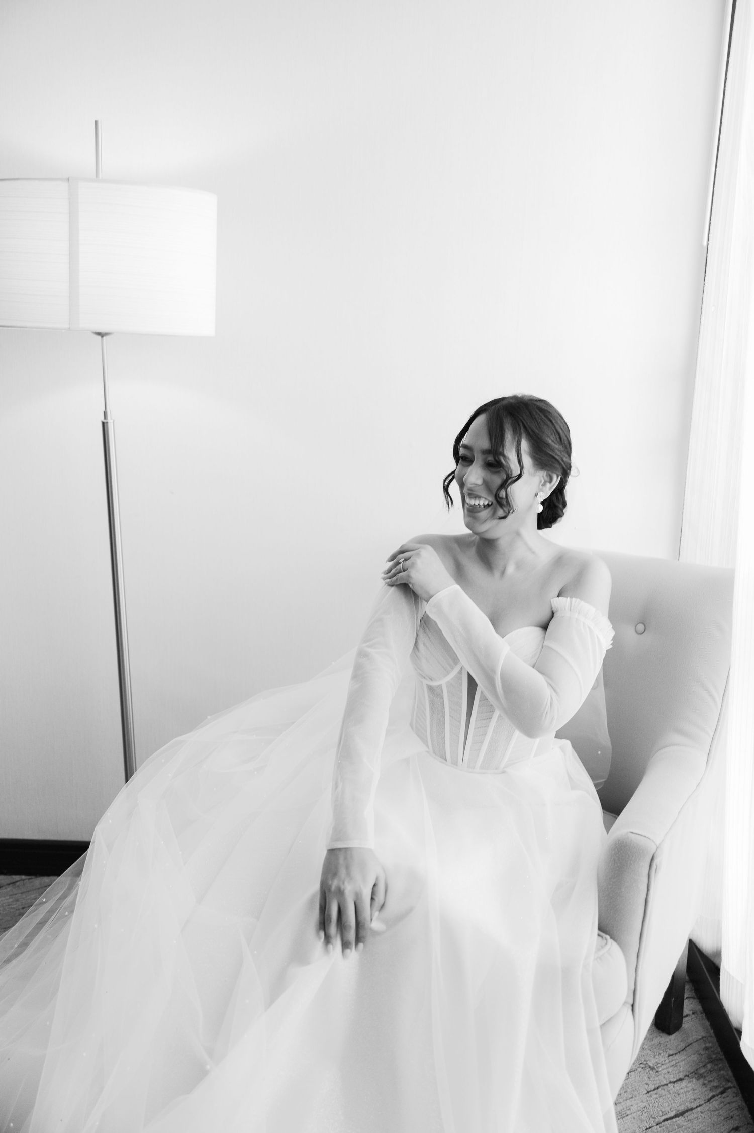 A bride in a flowing white wedding gown sits gracefully on a white couch during her wedding preparations.