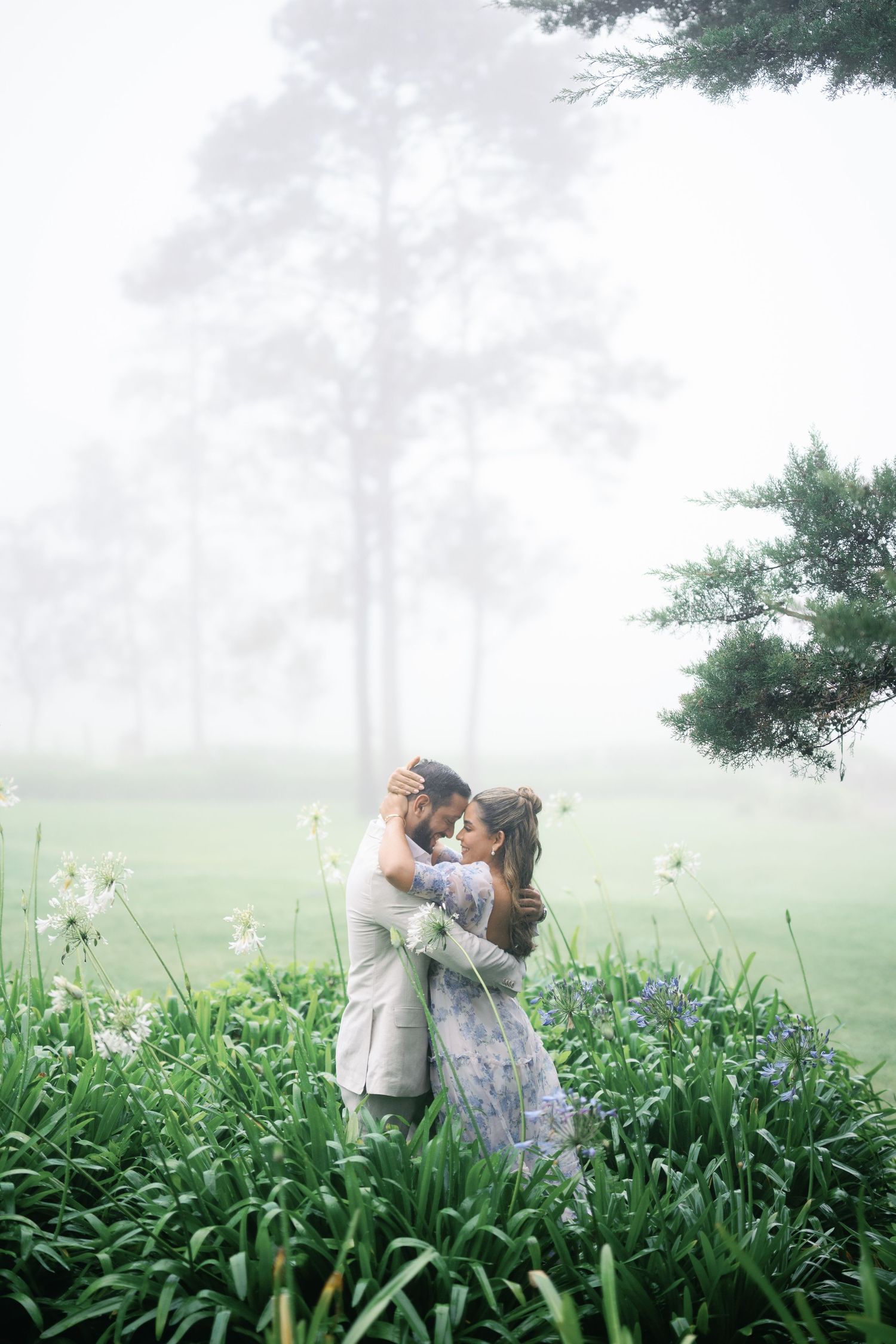 A couple shares a romantic kiss in a misty meadow surrounded by tall grass and wildflowers on a foggy morning.