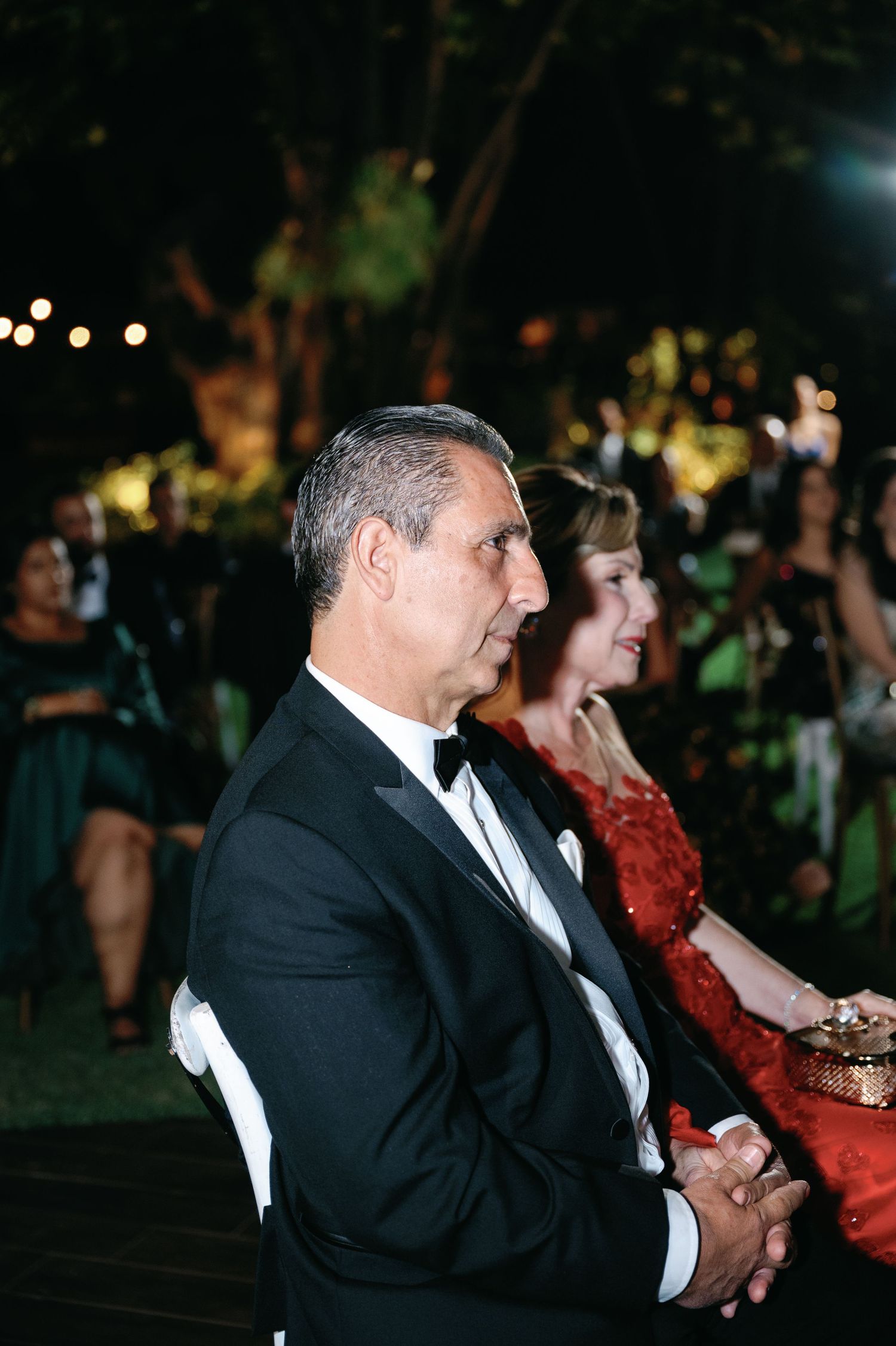 A couple in formal attire watches an evening outdoor event, with the man in a tuxedo and woman in a red dress.