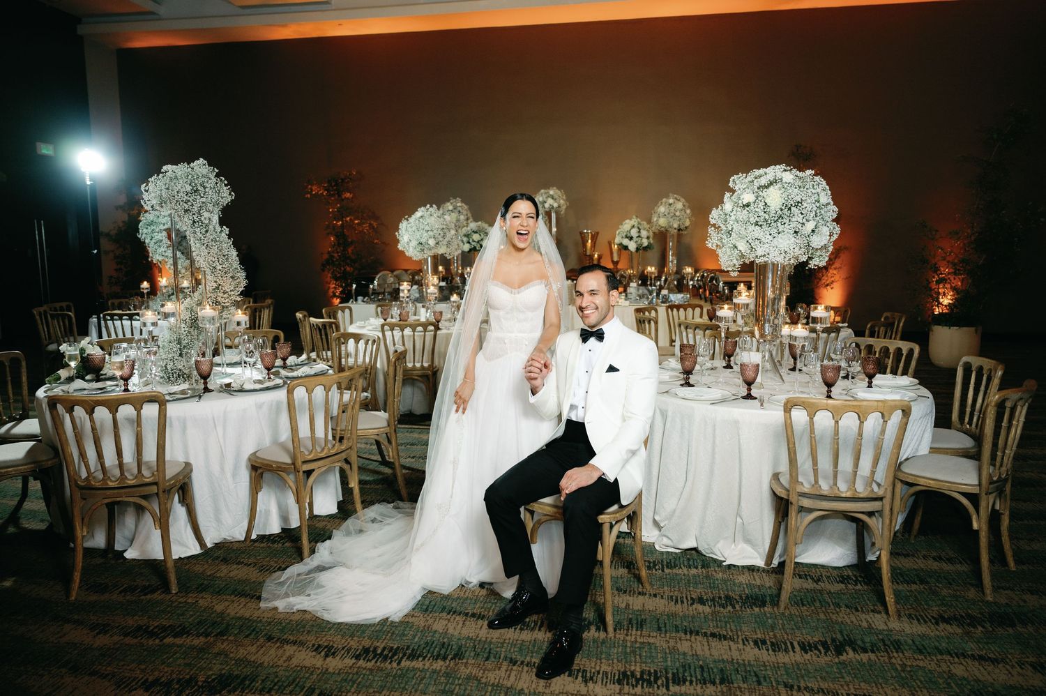 Wedding reception venue decorated with elegant white floral centerpieces and chiavari chairs in a romantic evening setting.