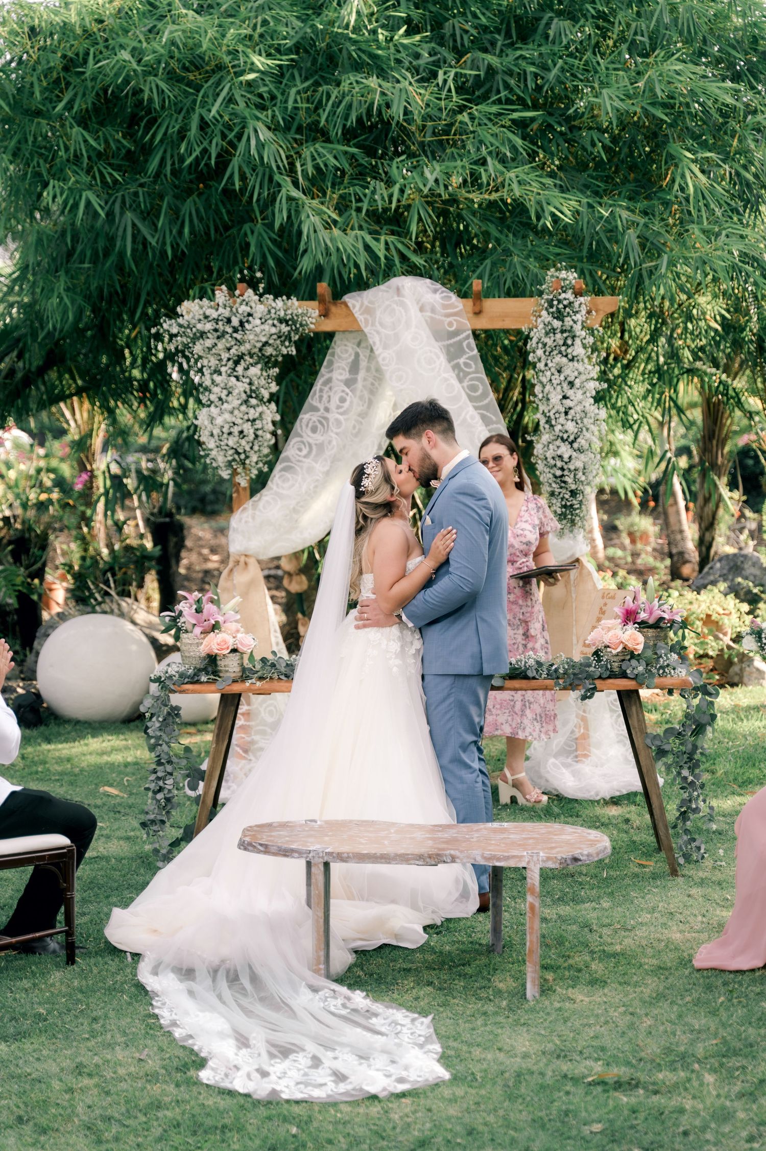 A romantic outdoor wedding ceremony under a wooden arch decorated with white floral arrangements and flowing fabric.