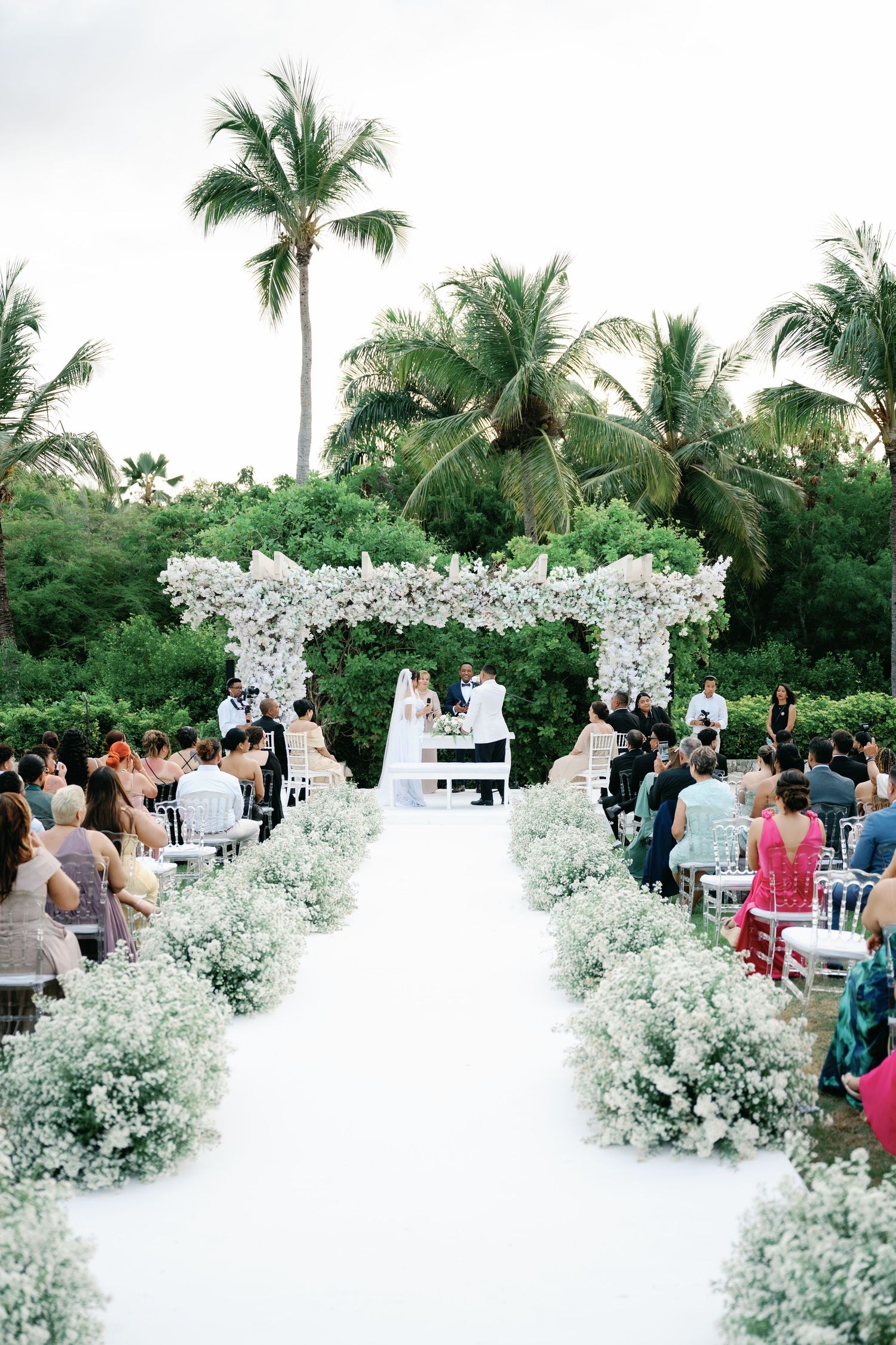Outdoor tropical wedding ceremony setup with white floral arrangements and palm trees in the background.
