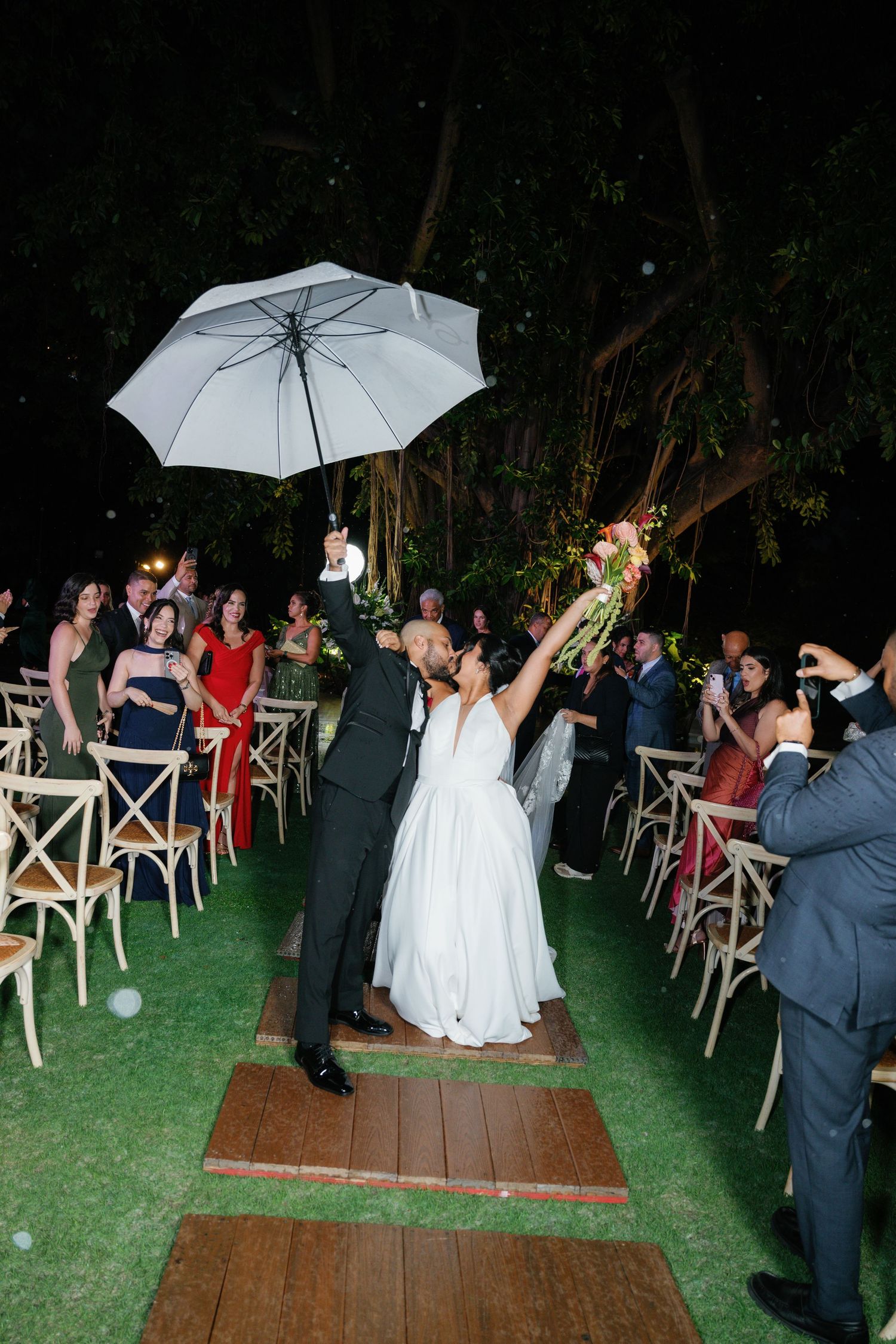 A couple celebrates at their outdoor wedding ceremony under a white umbrella while guests look on from wooden chairs.