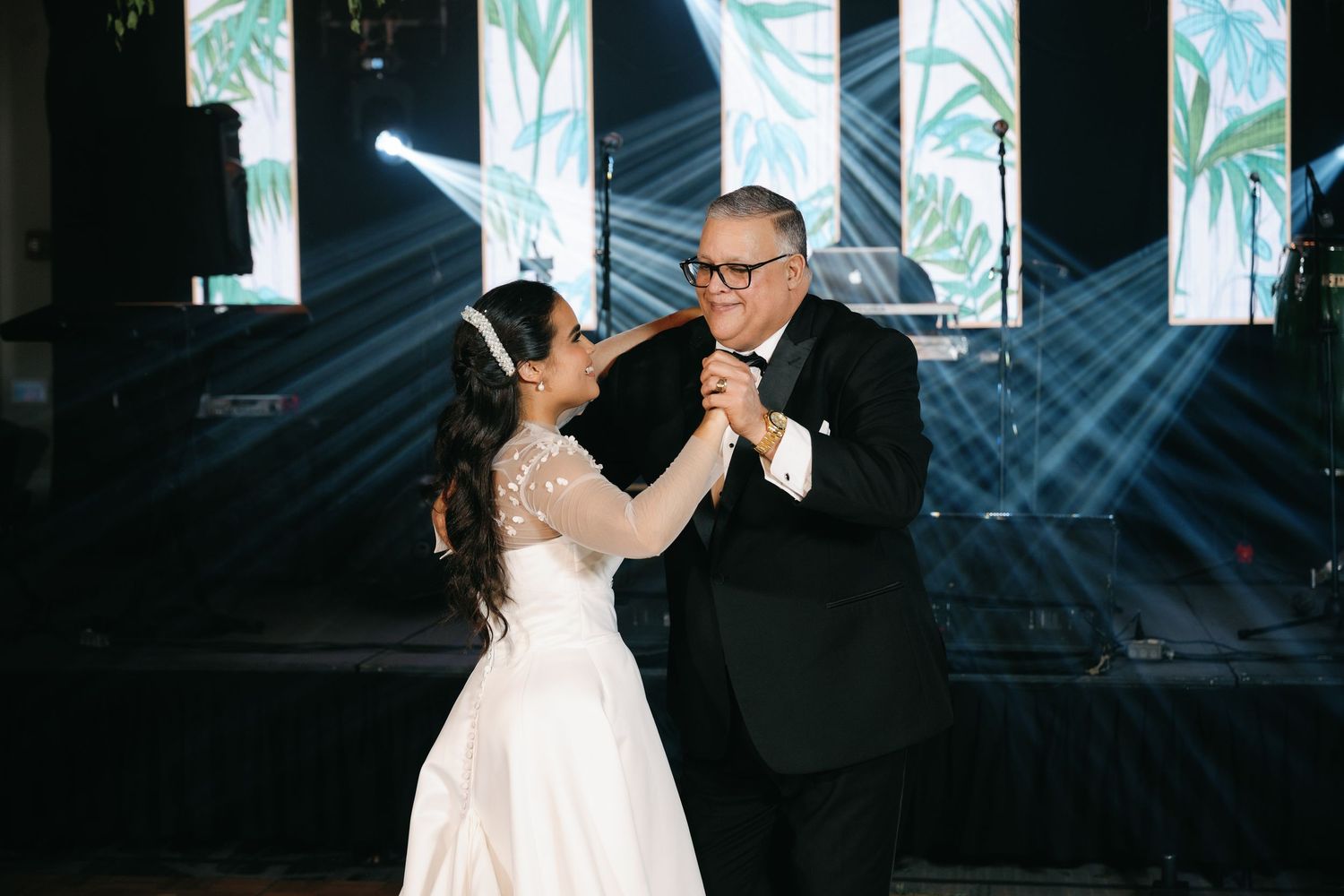 A couple dances together in a dark ballroom with dramatic lighting and tall windows in the background.