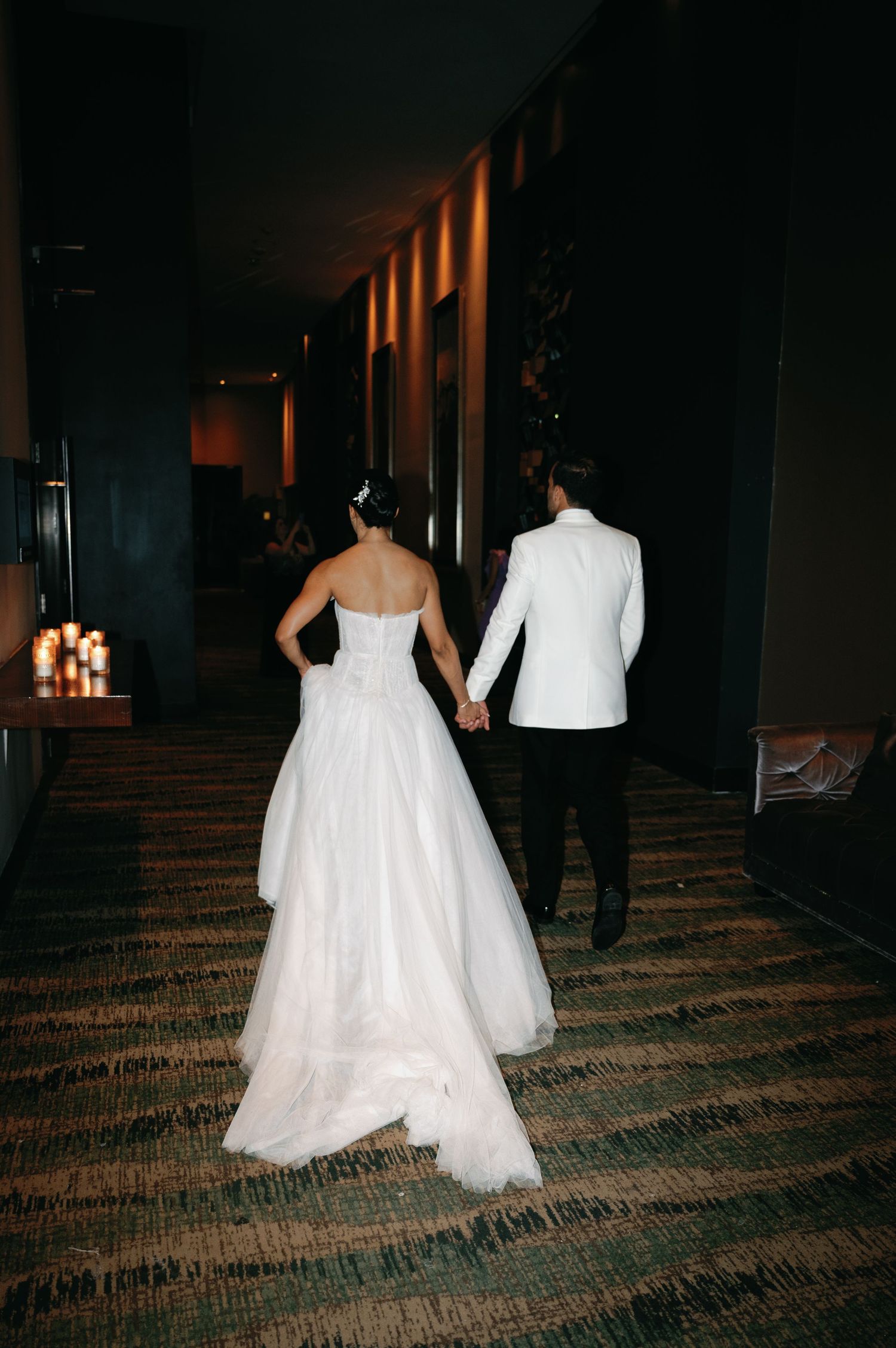 A couple in wedding attire walks down a dimly lit hotel hallway with patterned carpeting.