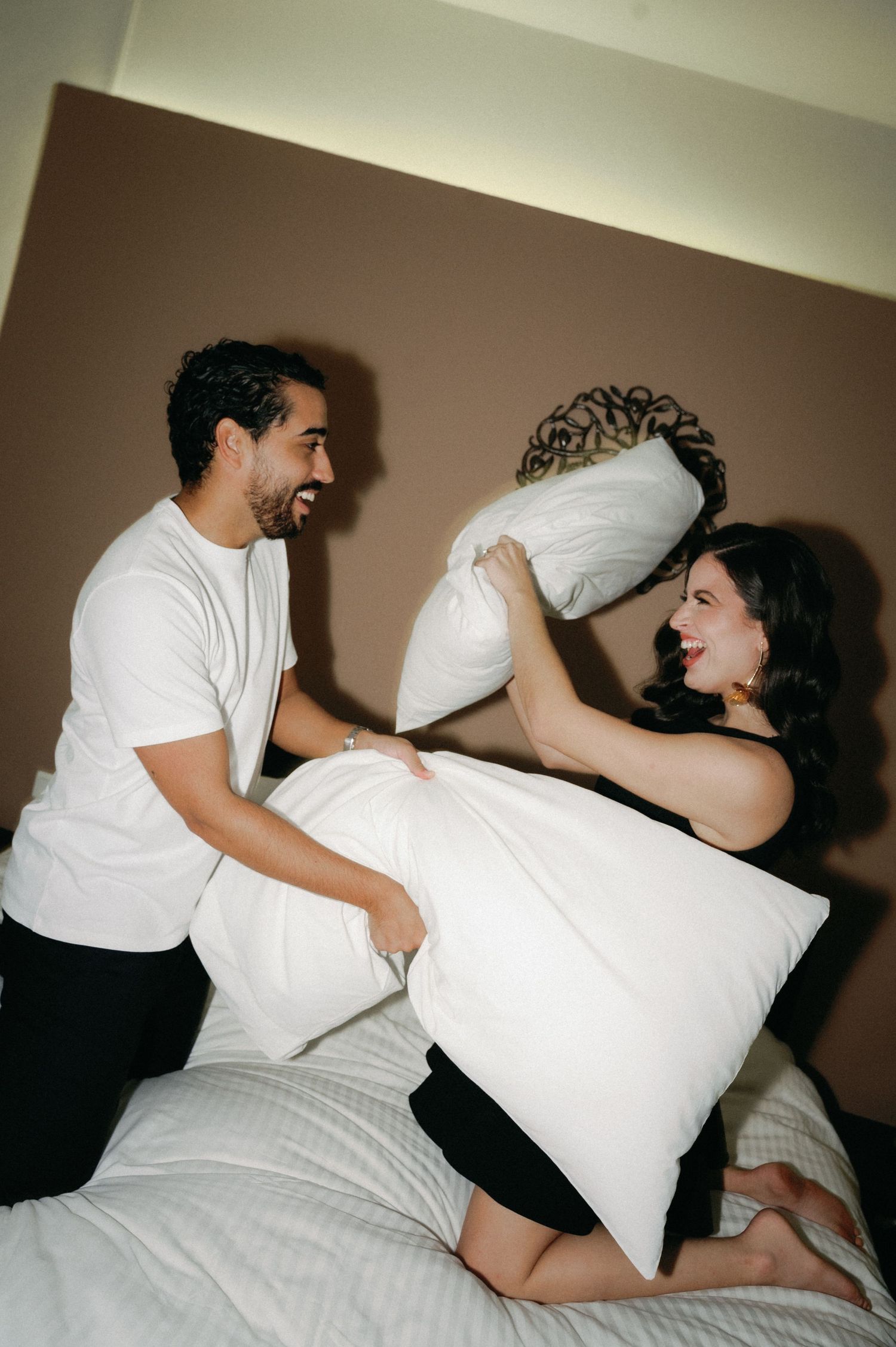 Two people engage in a playful pillow fight sequence on a bed wearing white and black clothing.