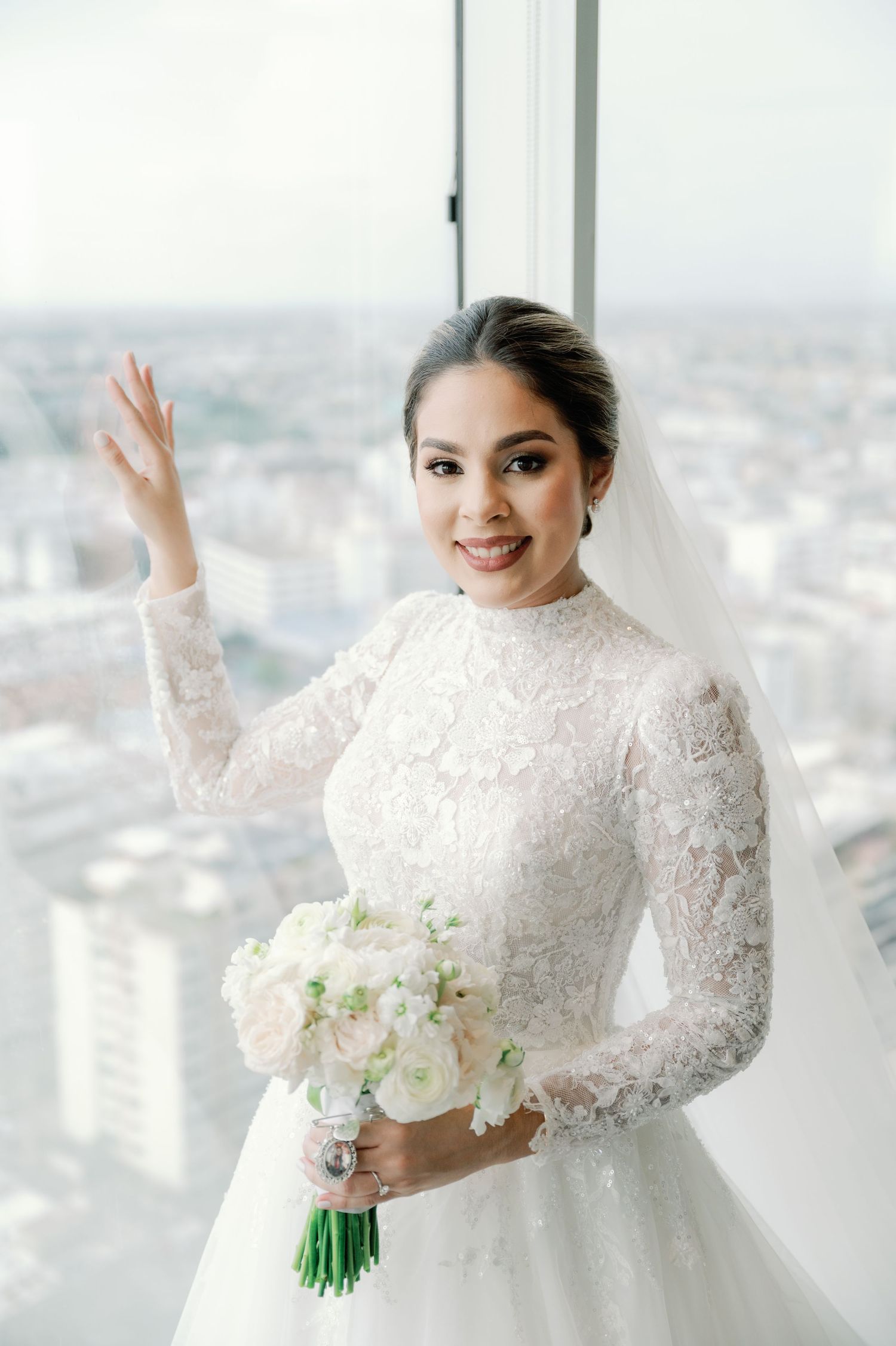 Elegant bride in white lace long-sleeve wedding dress holds white bouquet while posing near window with city view.