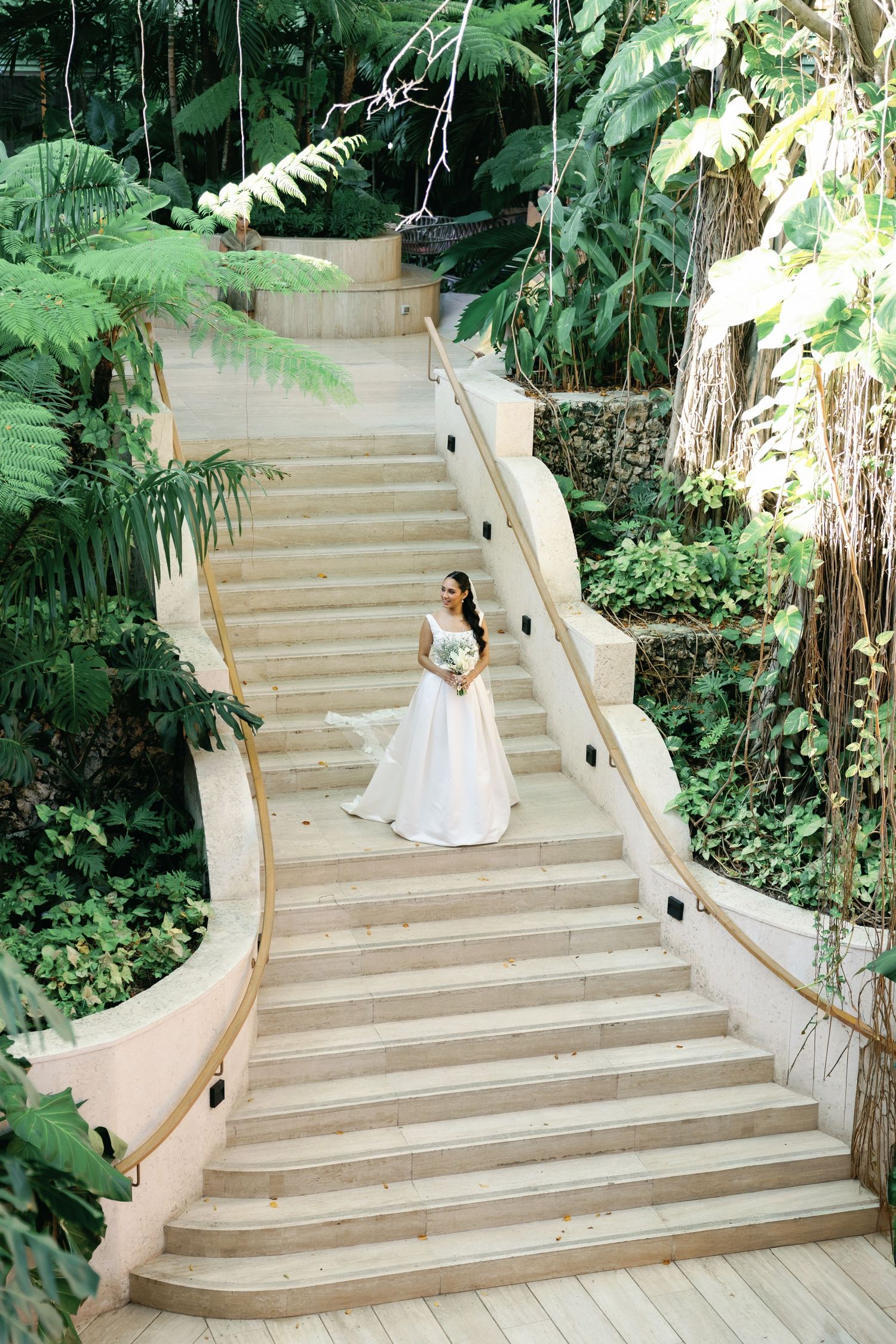 Bride in flowing white gown descends grand outdoor stone staircase surrounded by lush tropical greenery.
