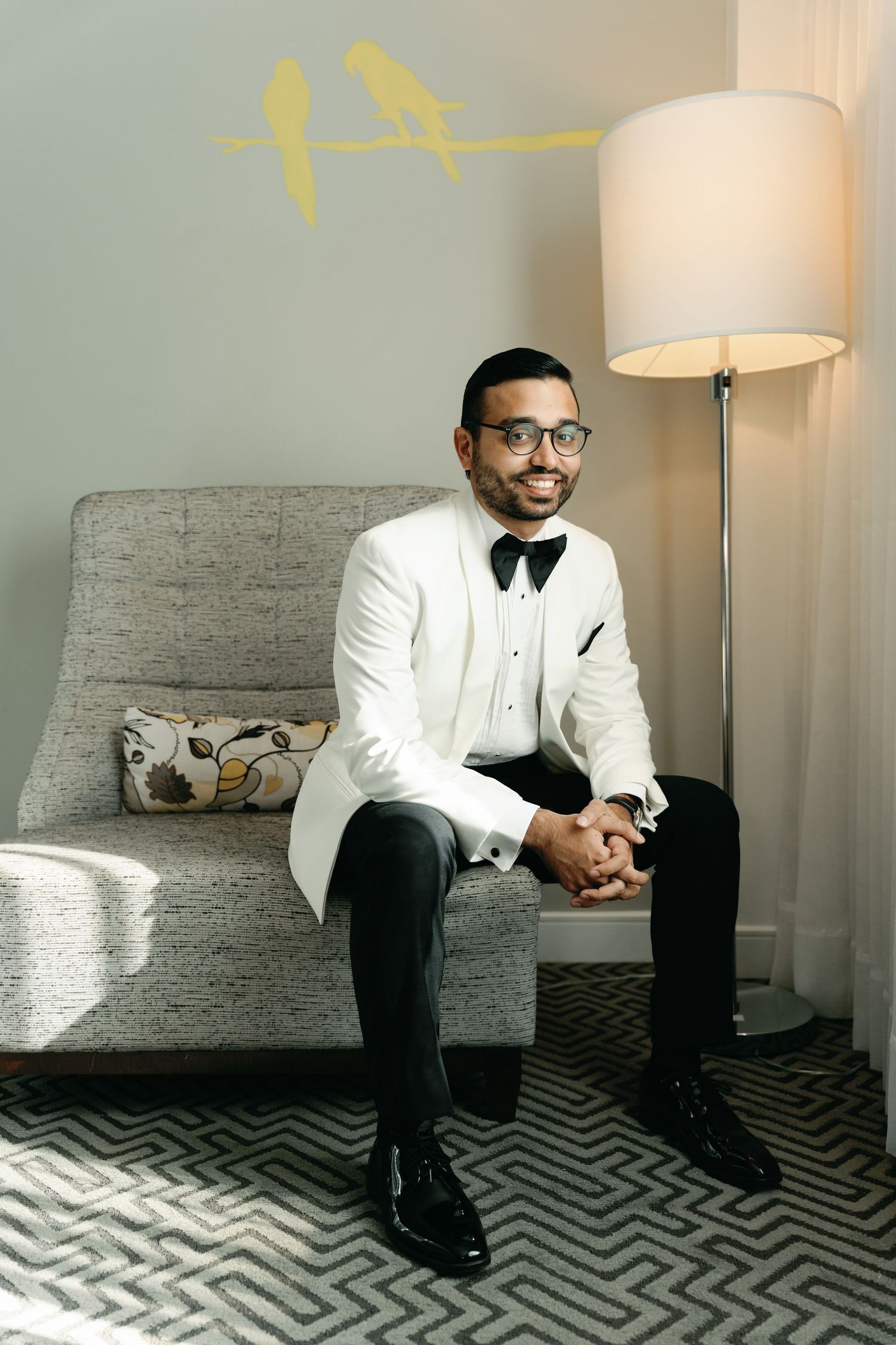 A person in a white tuxedo jacket and bow tie sits contemplatively on a gray couch with chevron-patterned flooring.
