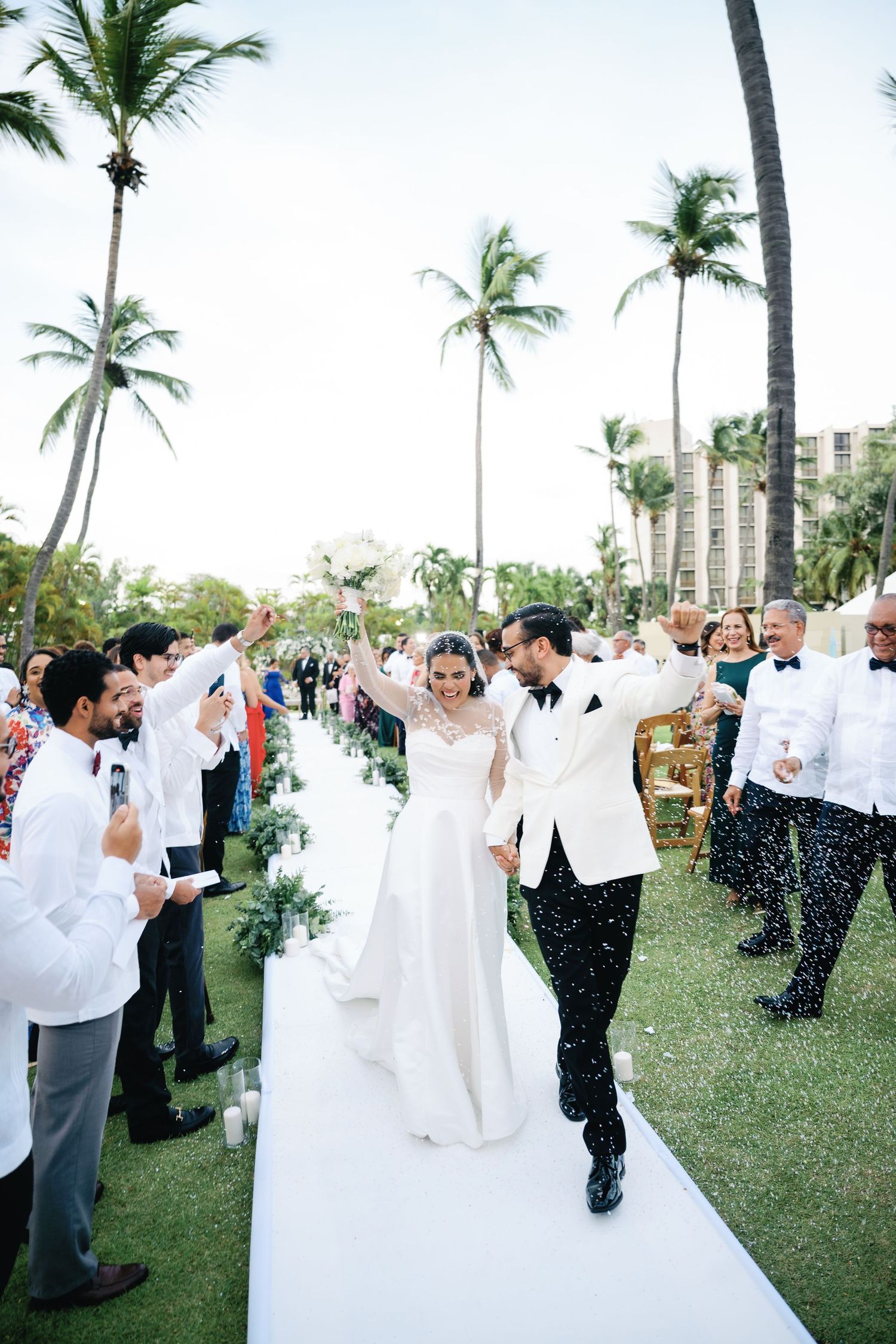 Wedding guests throw white flower petals at a tropical beach celebration with palm trees and ocean views.