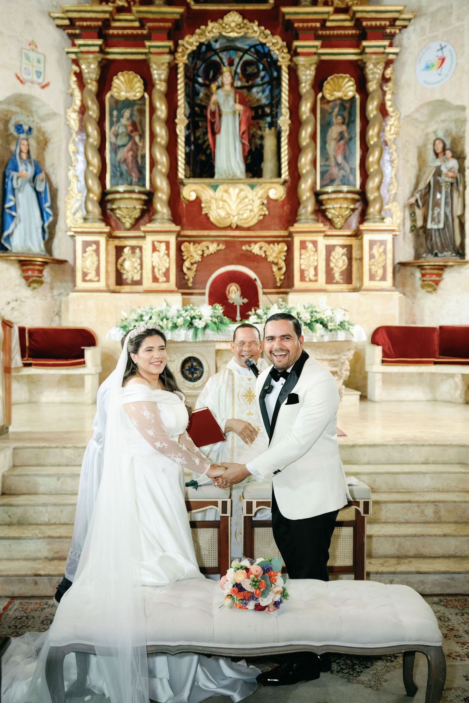 Wedding couple poses playfully on church altar steps with ornate religious artwork and statues in background.