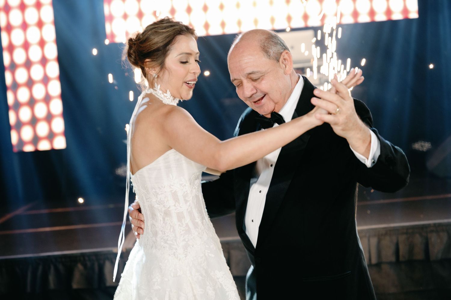 Wedding couple dances together in an elegant ballroom with dramatic lighting and spotlights.