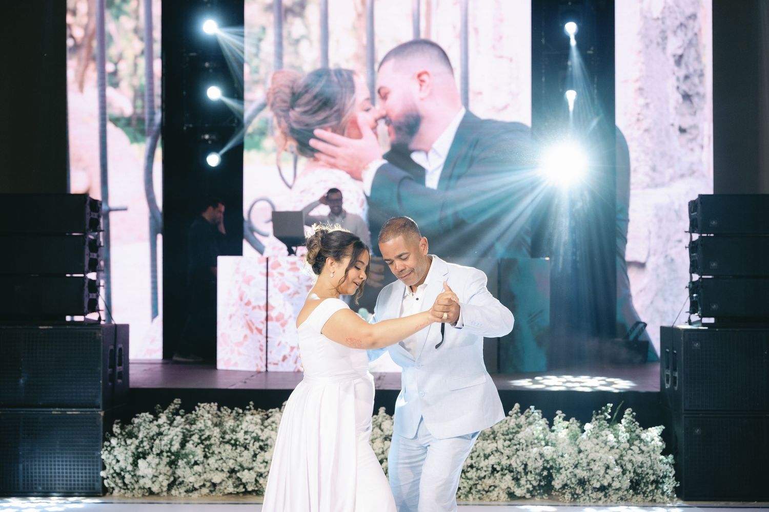 A couple shares their first dance at a wedding reception surrounded by white floral arrangements and dramatic lighting.