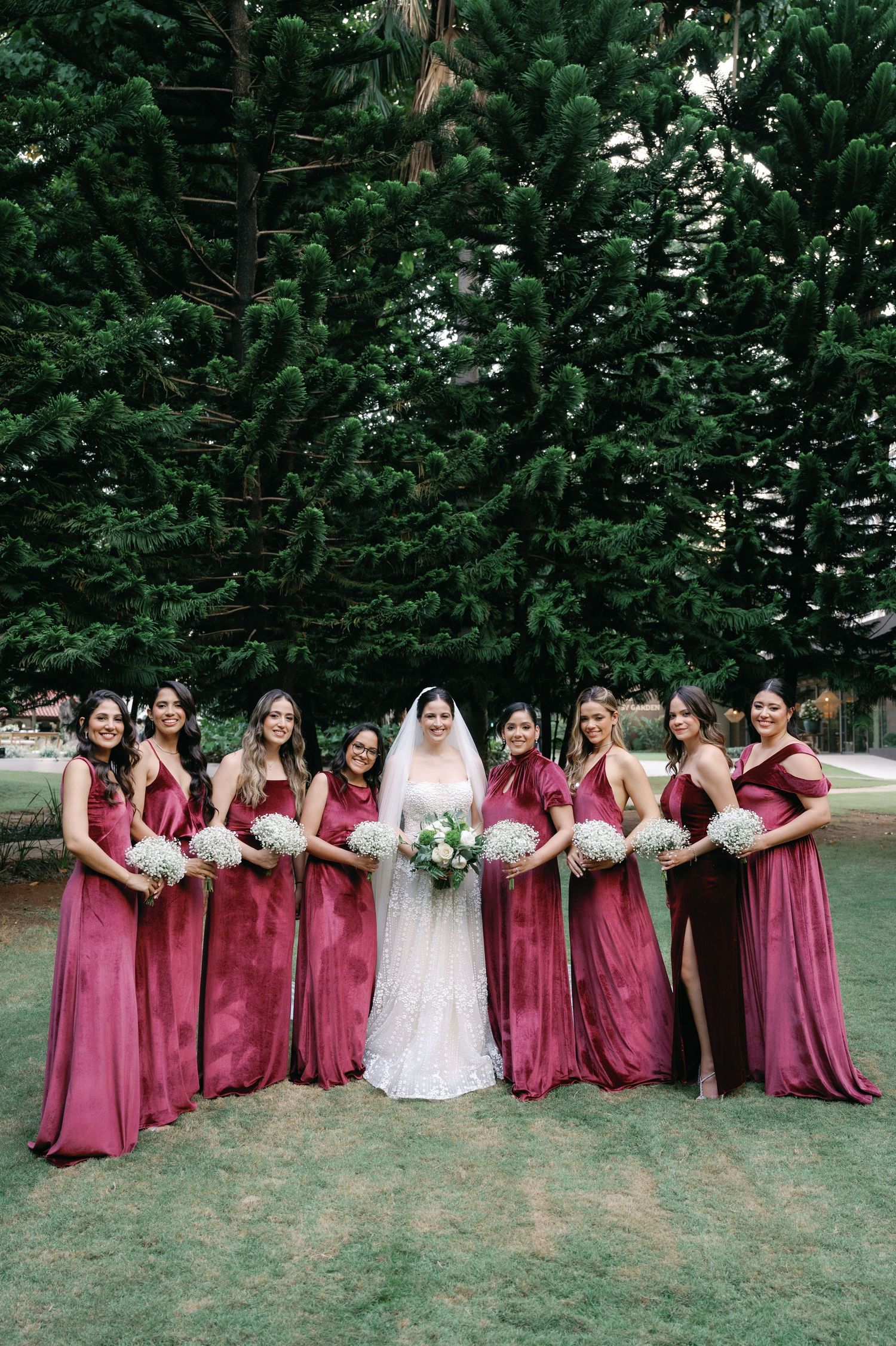 Bridesmaids in burgundy dresses pose in a line with the bride on a grassy lawn surrounded by evergreen trees.