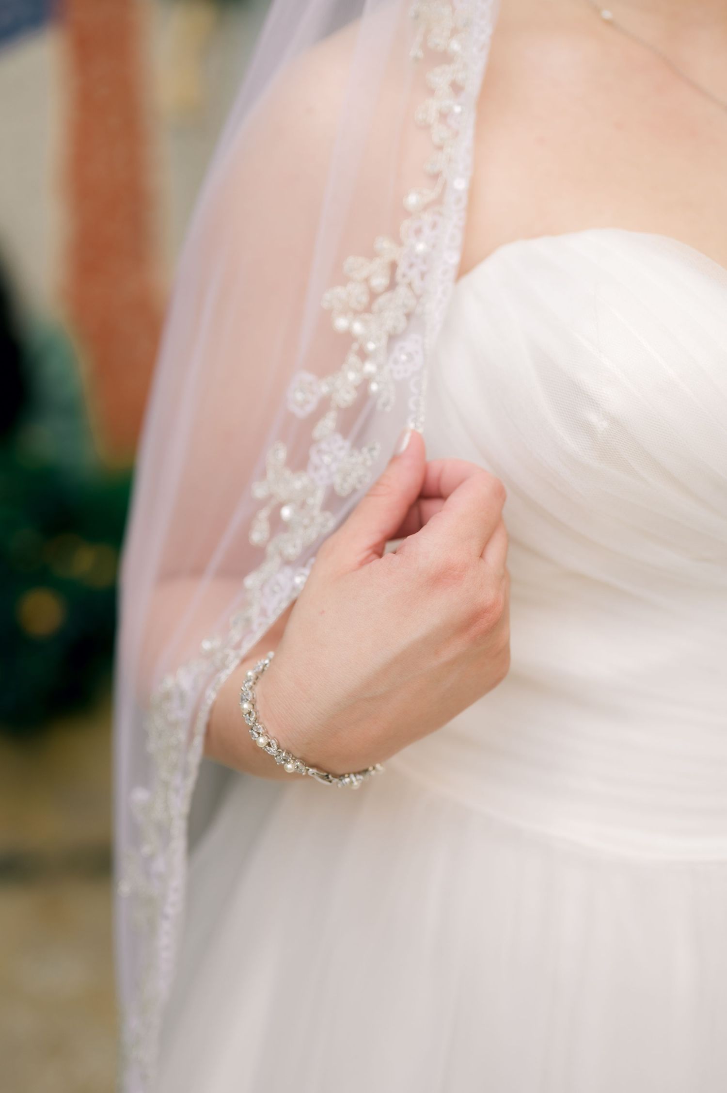 A delicate white bridal veil with lace detailing is gently held against a wedding dress.