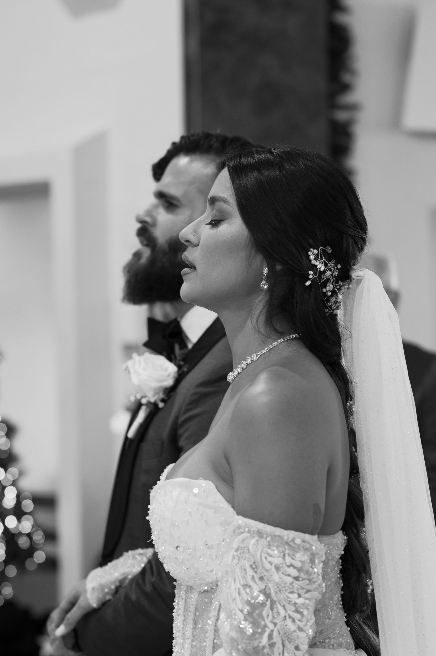 A black and white wedding photo showing a bride in a lace gown and veil standing beside her groom in a dark suit.