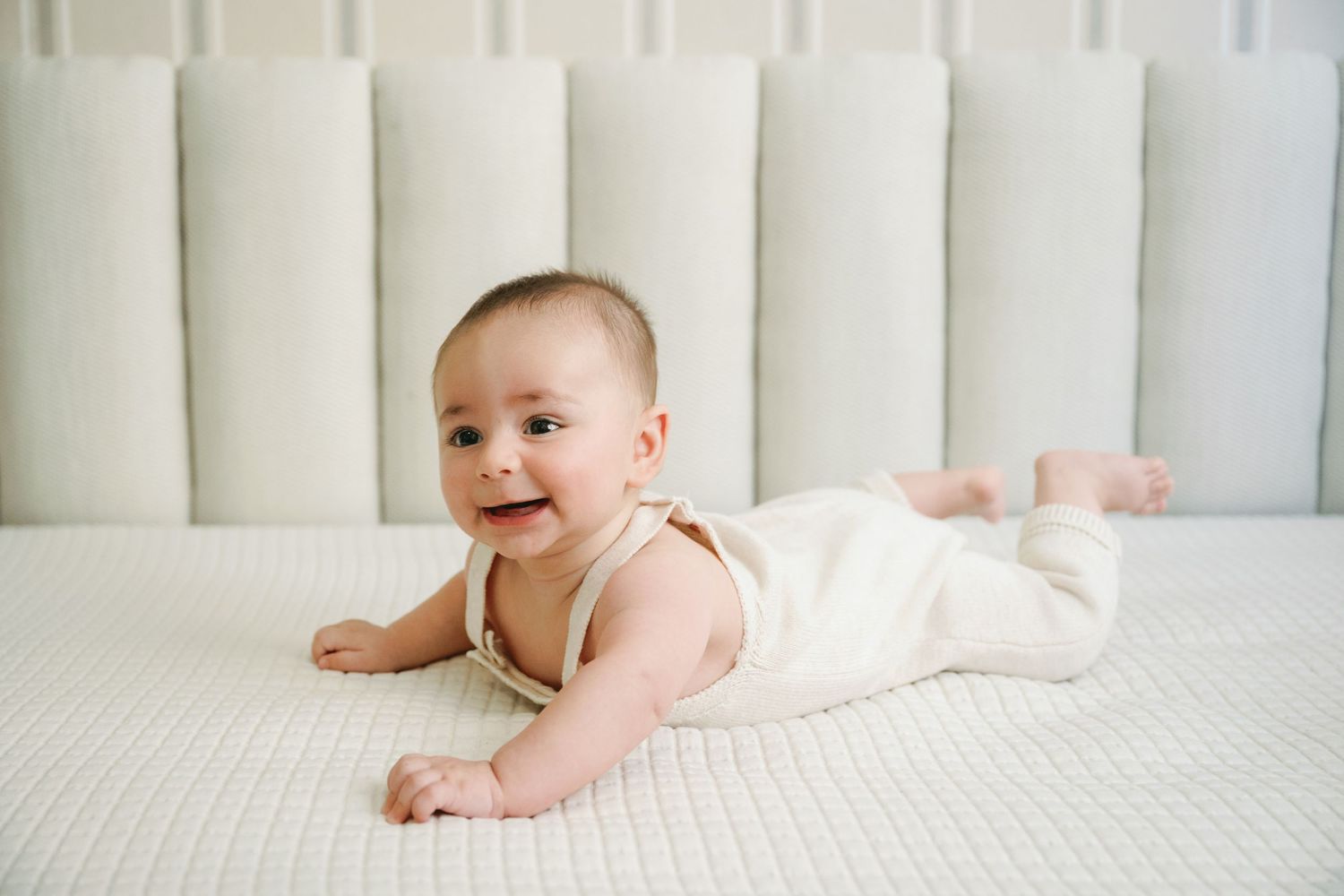 Smiling baby in white romper doing tummy time on a white mattress with a padded headboard background.