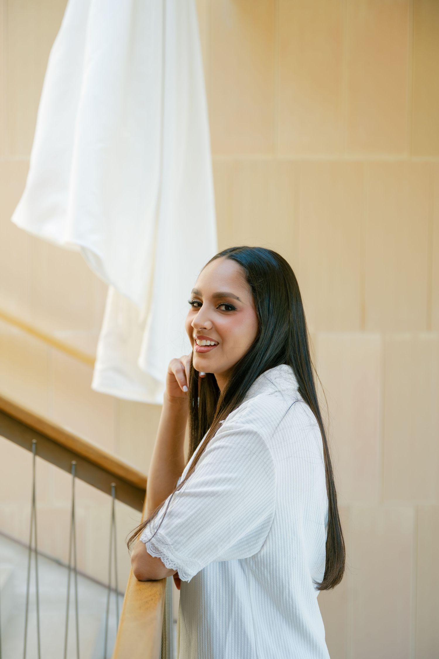 A person in a white button-down shirt smiles while standing near a staircase railing with soft lighting in the background.