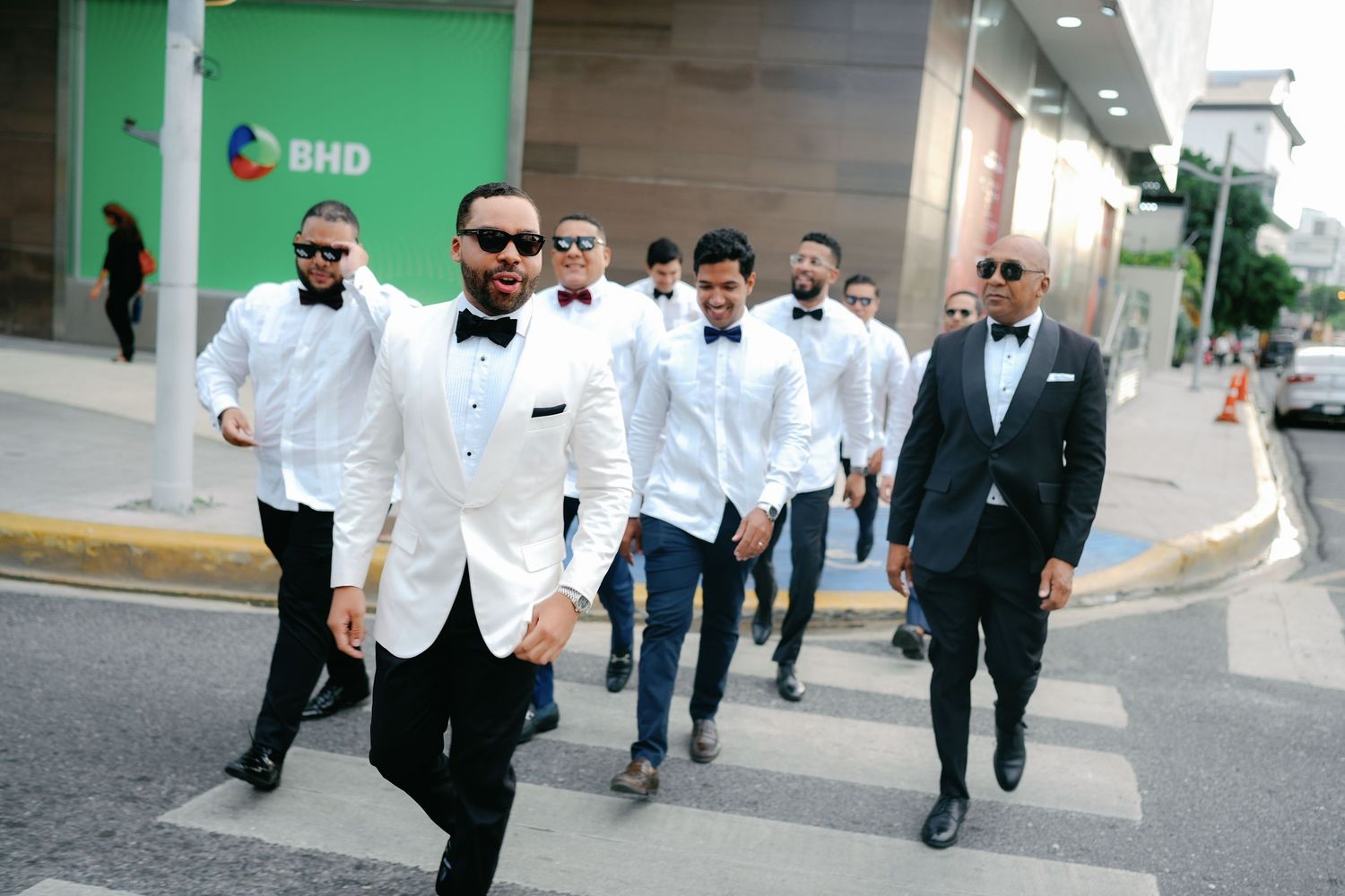 A group of men in white jackets and suits walk confidently down a city street in formation.