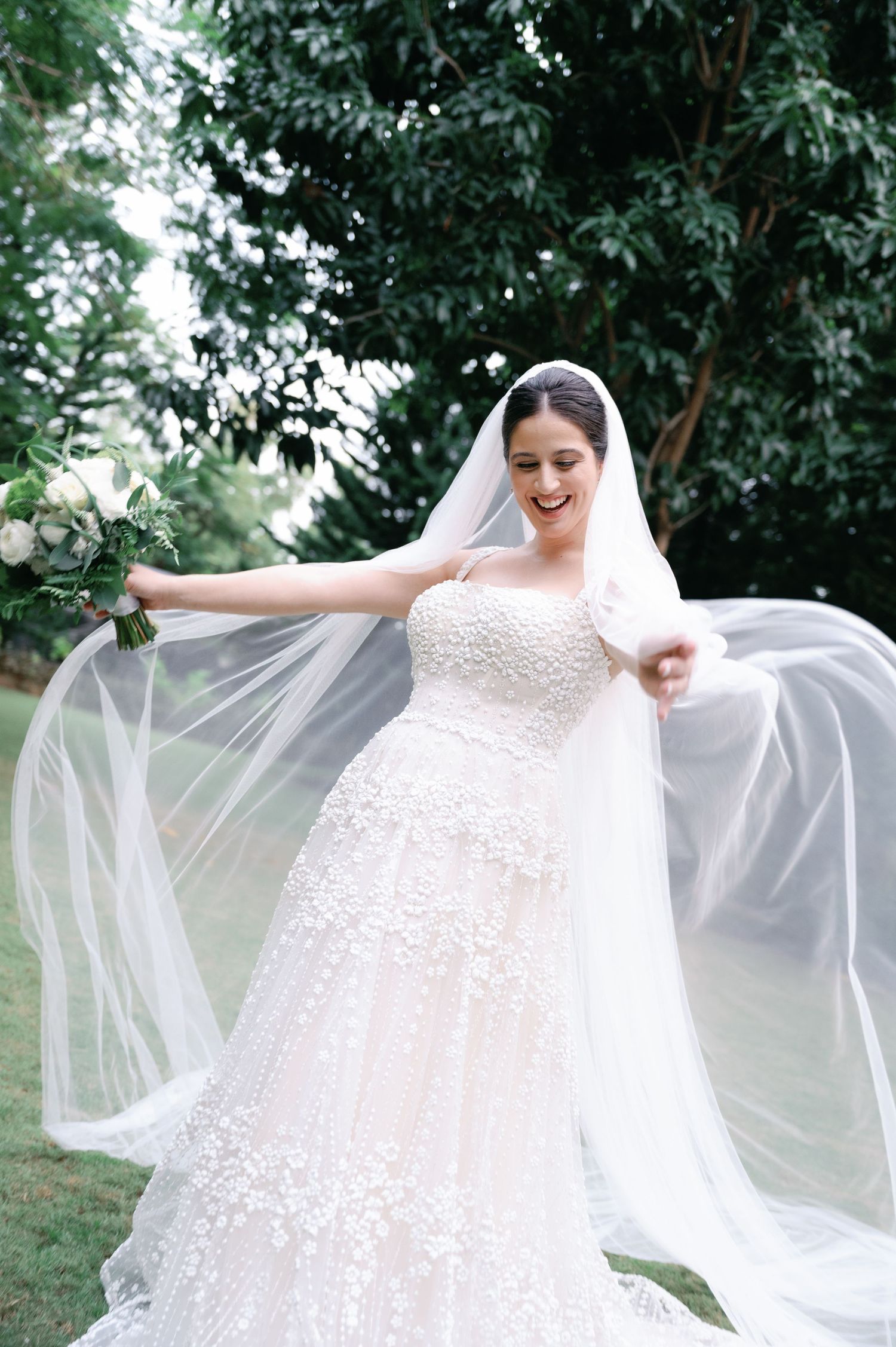 A bride in a white lace wedding dress twirls joyfully outdoors, making her veil float gracefully in the breeze.