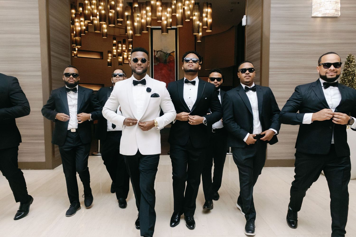 A panoramic group photo of groomsmen in black suits and sunglasses posing together at a wedding venue.