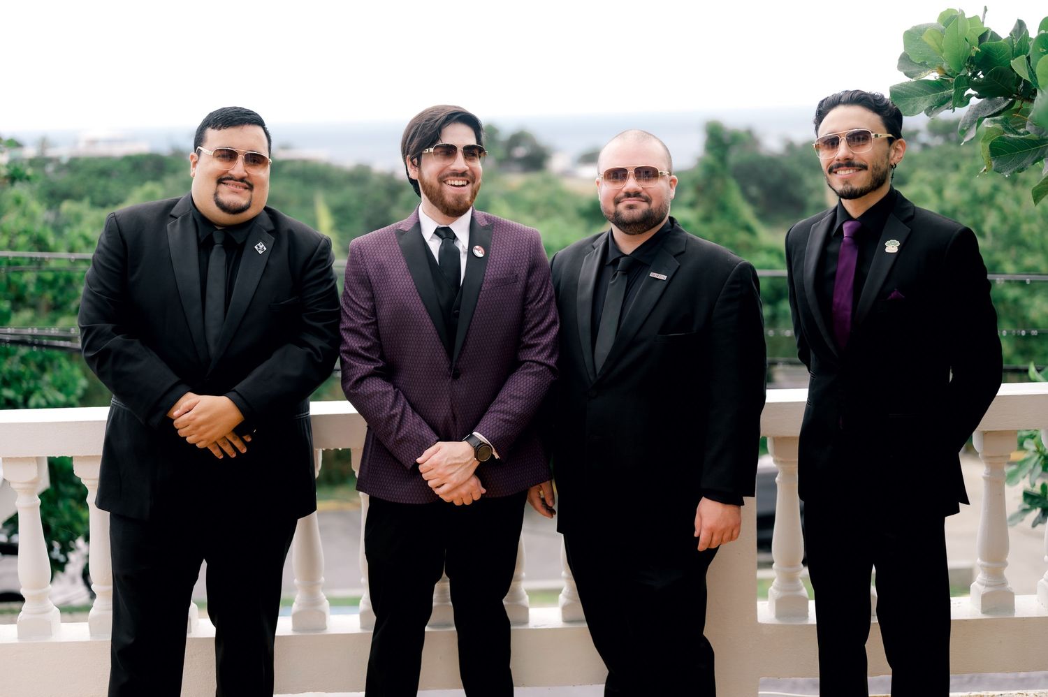 A group of men in formal black attire and burgundy suits pose together outdoors for a wedding party photo.
