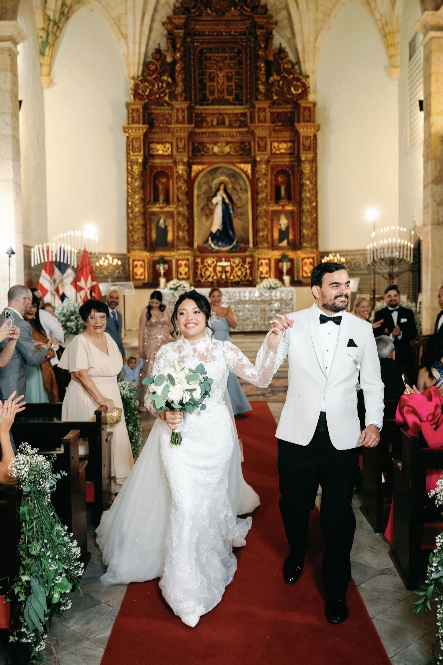 Wedding ceremony in an ornate church with white floral decorations and guests lining the aisle during the recessional.