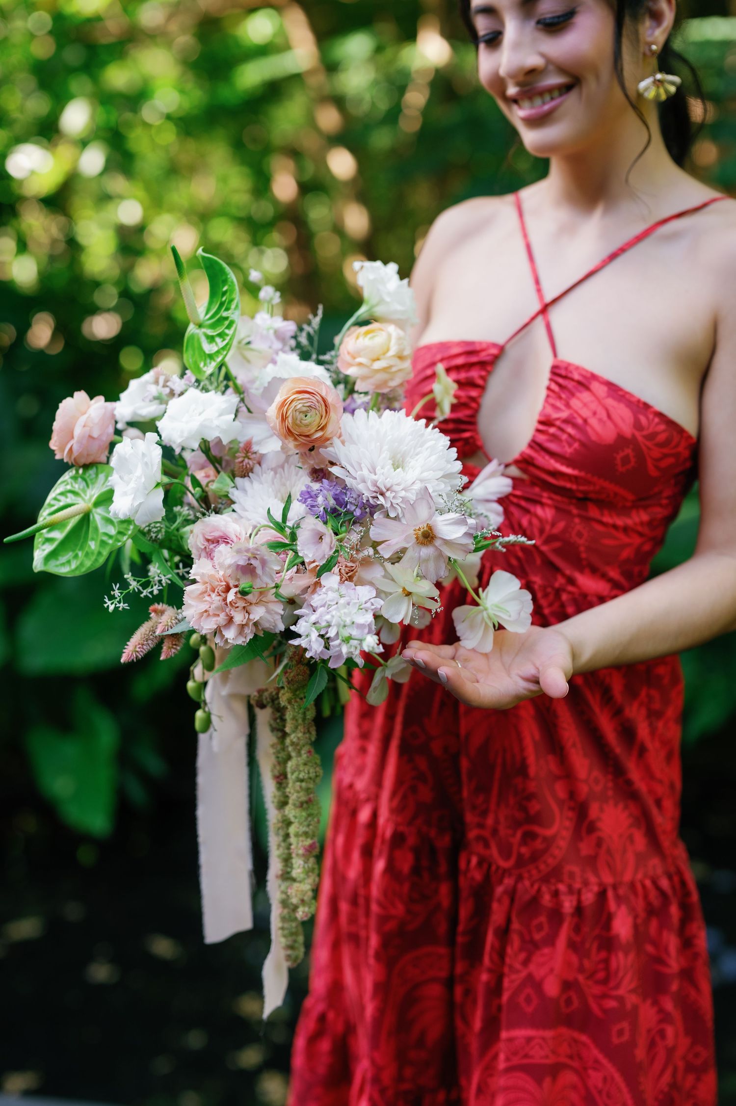 A person in a red velvet dress holds a delicate bouquet of white and pink flowers against a green garden backdrop.