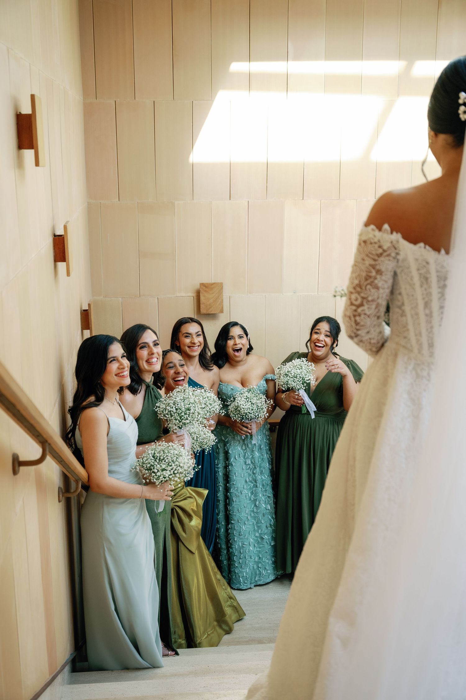 Bridesmaids in mint green and olive dresses gather around a white wedding gown hanging in a bridal suite.