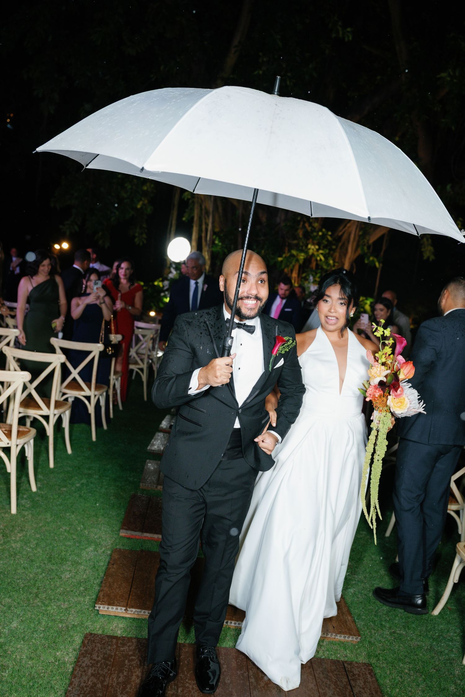 Wedding couple shares white umbrellas during outdoor rainy evening reception on grass lawn with guests nearby.
