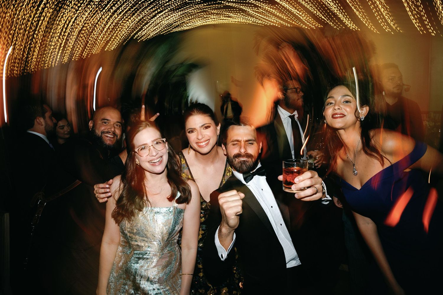 Formal party guests raise glasses in celebration under a decorative ceiling of twinkling lights.