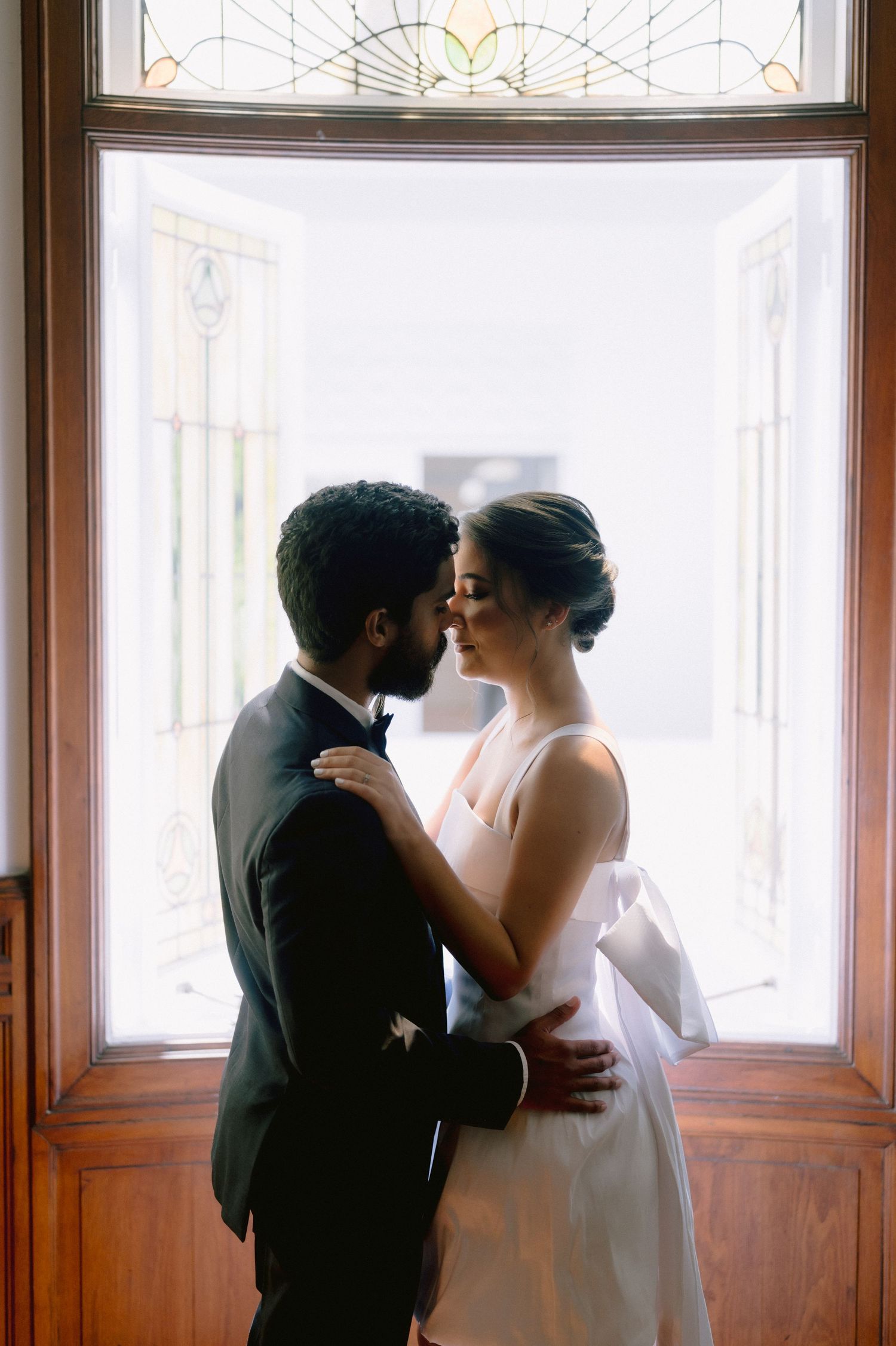 A romantic moment between newlyweds sharing a kiss by a window with decorative stained glass details.