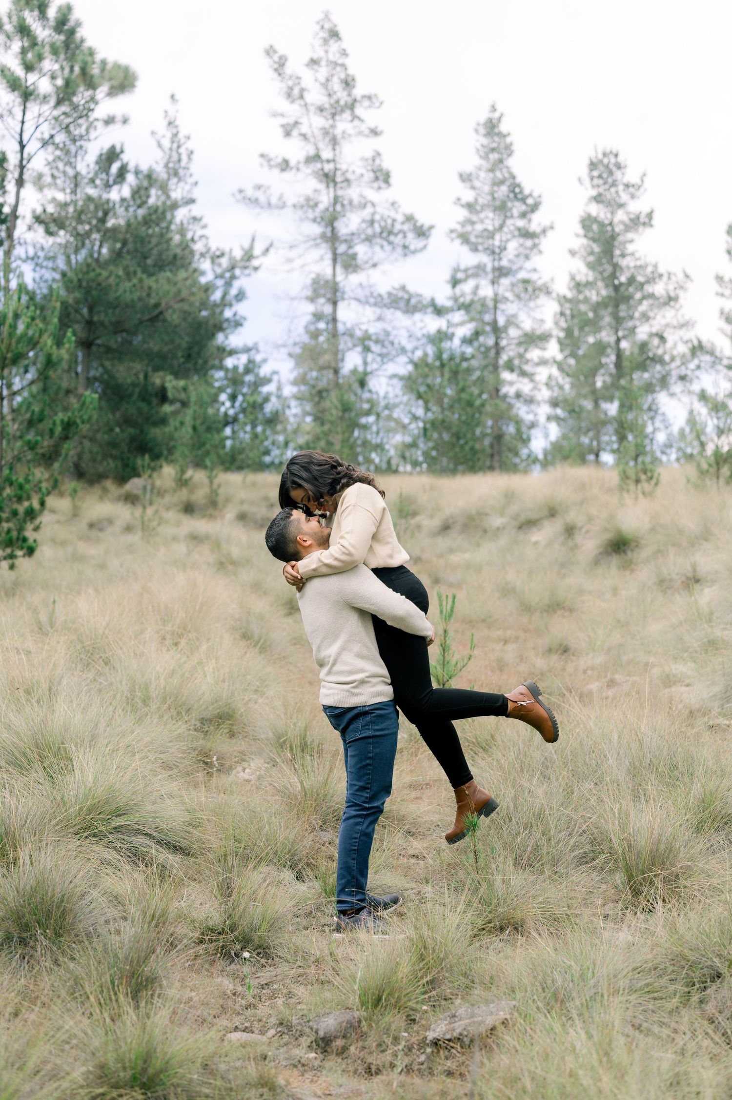 A romantic couple embraces and dances together in a field of tall grass with pine trees in the background.